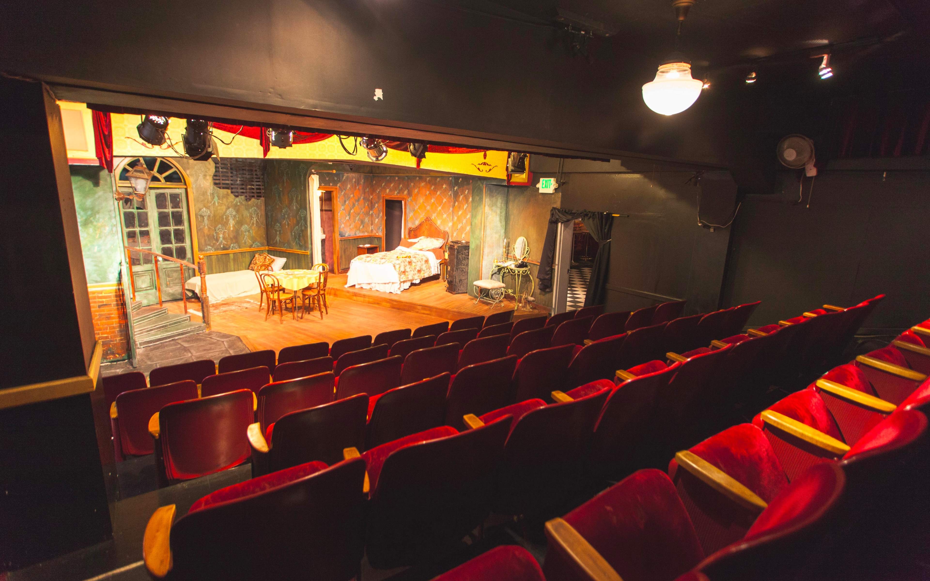 An empty theater with red upholstered seats facing a stage set with a bed, table, and various props in a detailed interior.