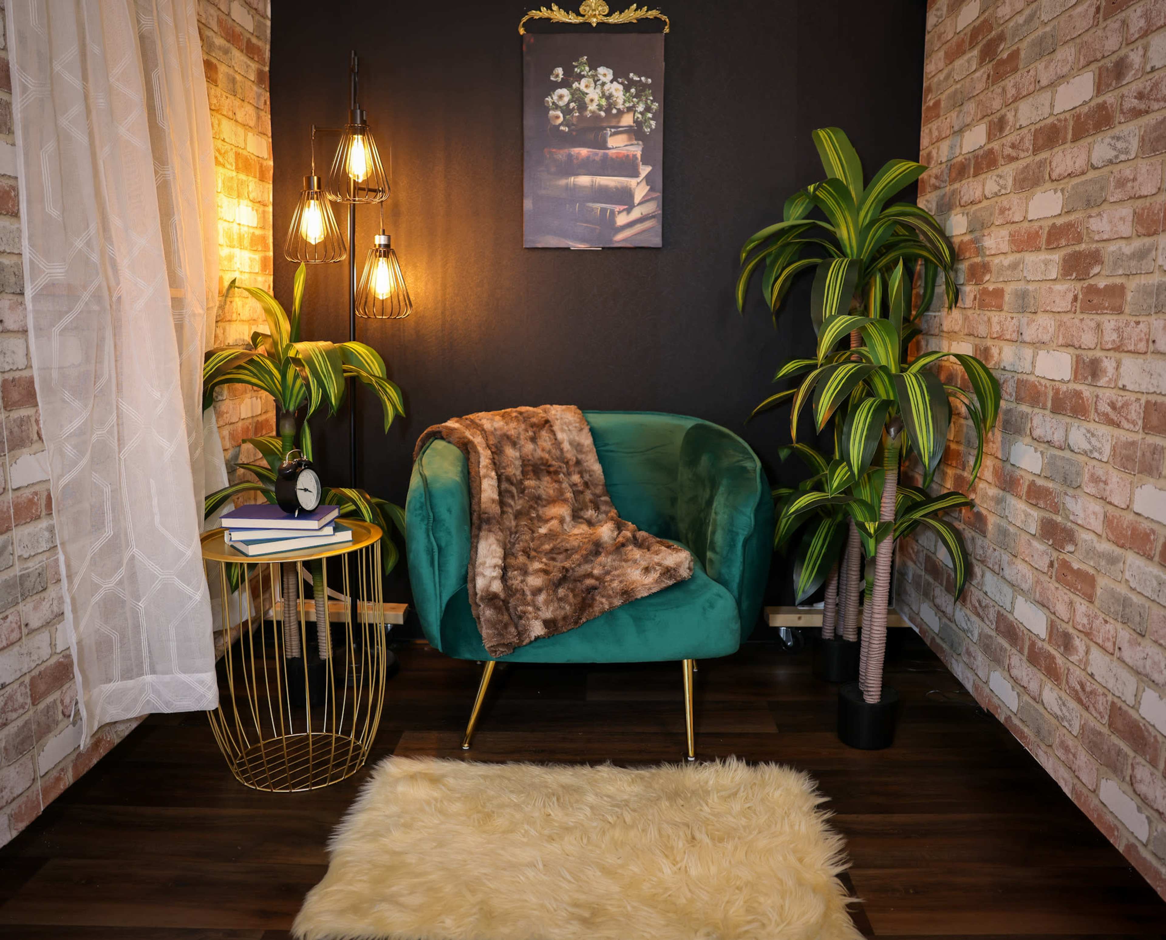 The image features a cozy corner with a green velvet chair, a round gold side table, a fluffy white rug, hanging pendant lights, and potted plants against a brick wall backdrop.