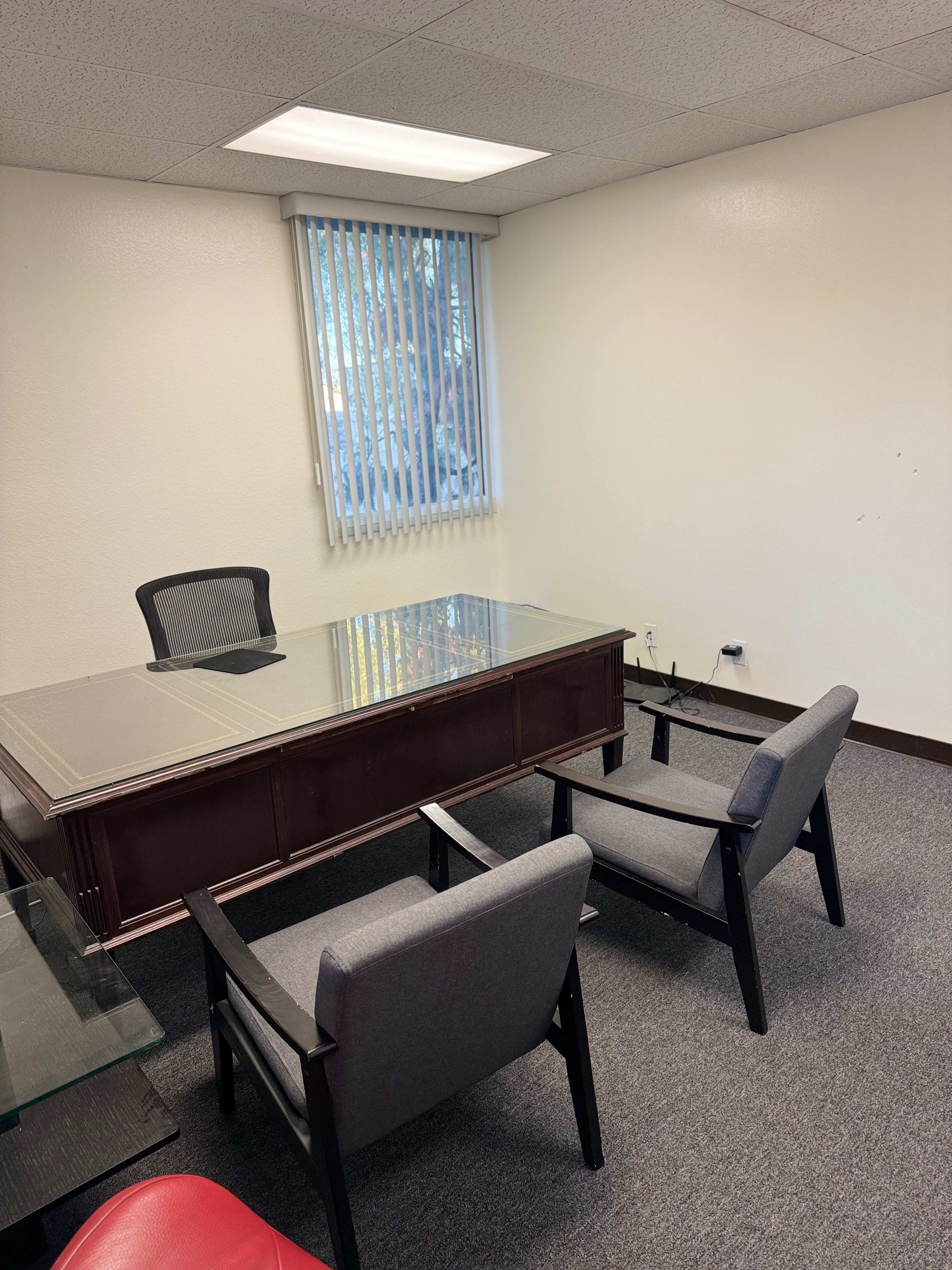 The image shows a small office with a large desk, two gray chairs in front of it, and vertical blinds on a window.