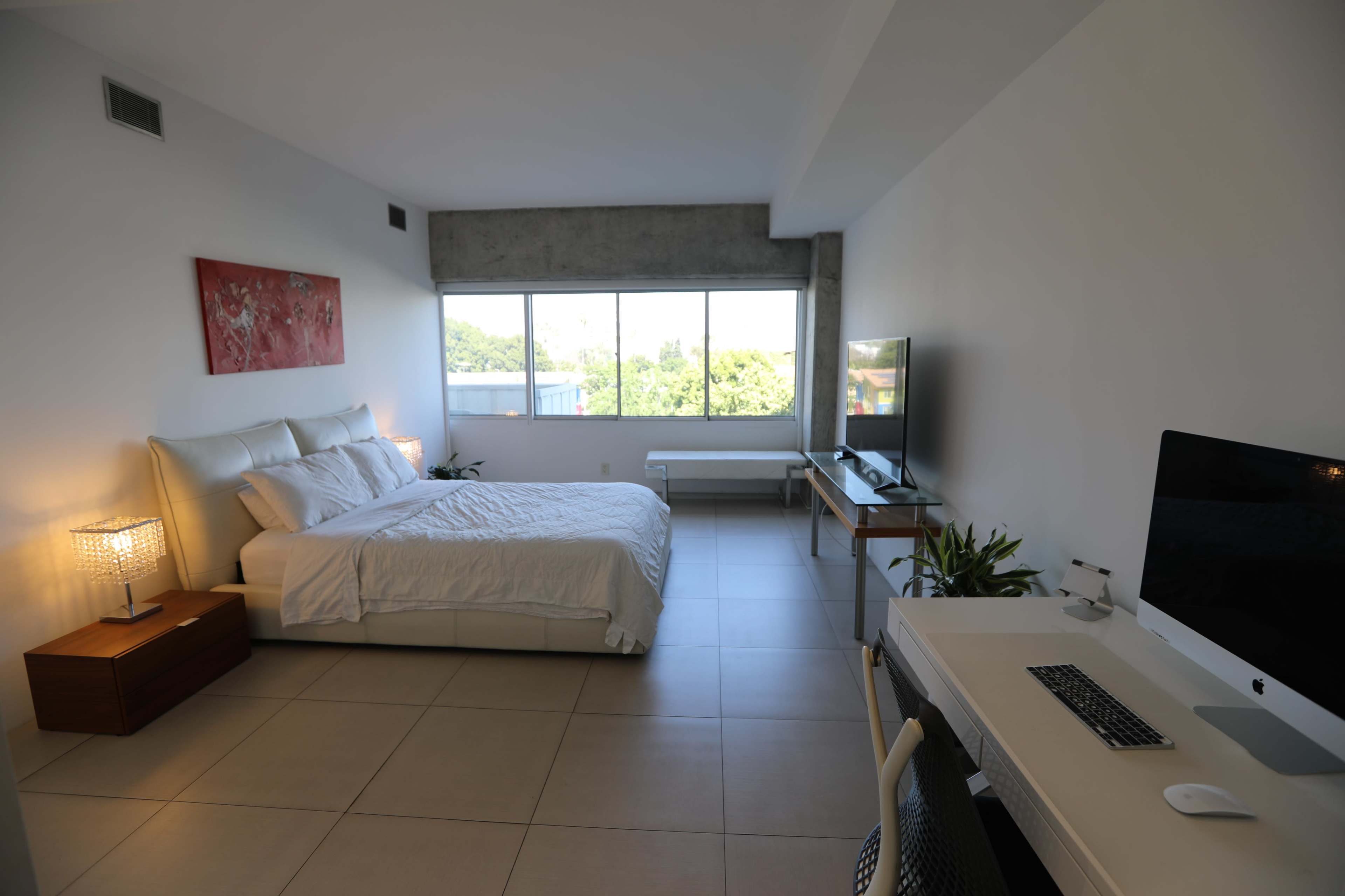 A modern bedroom features a bed with white linens, a desk with a computer, and large windows offering a view of greenery outside.