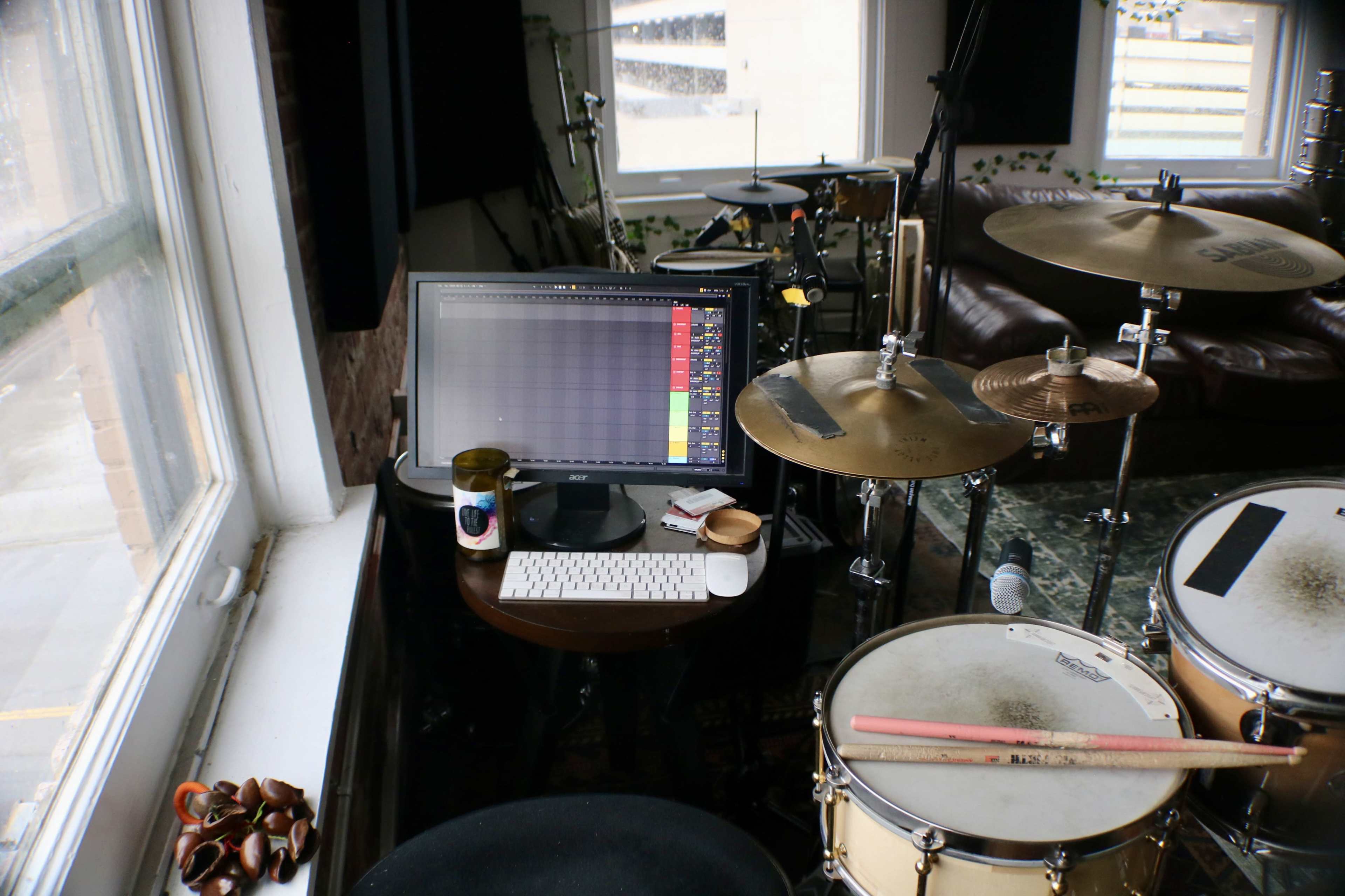 A computer monitor and keyboard are set on a table next to a drum kit in a sunny room with large windows.
