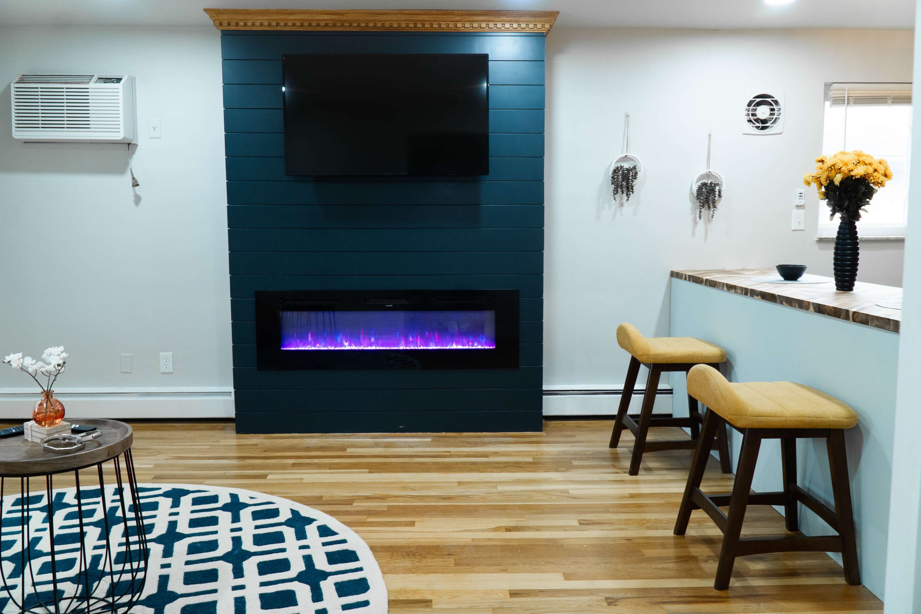 A modern living space with a blue accent wall featuring a flat-screen TV and an electric fireplace, accompanied by a round rug, a small table, and two bar stools at a counter.