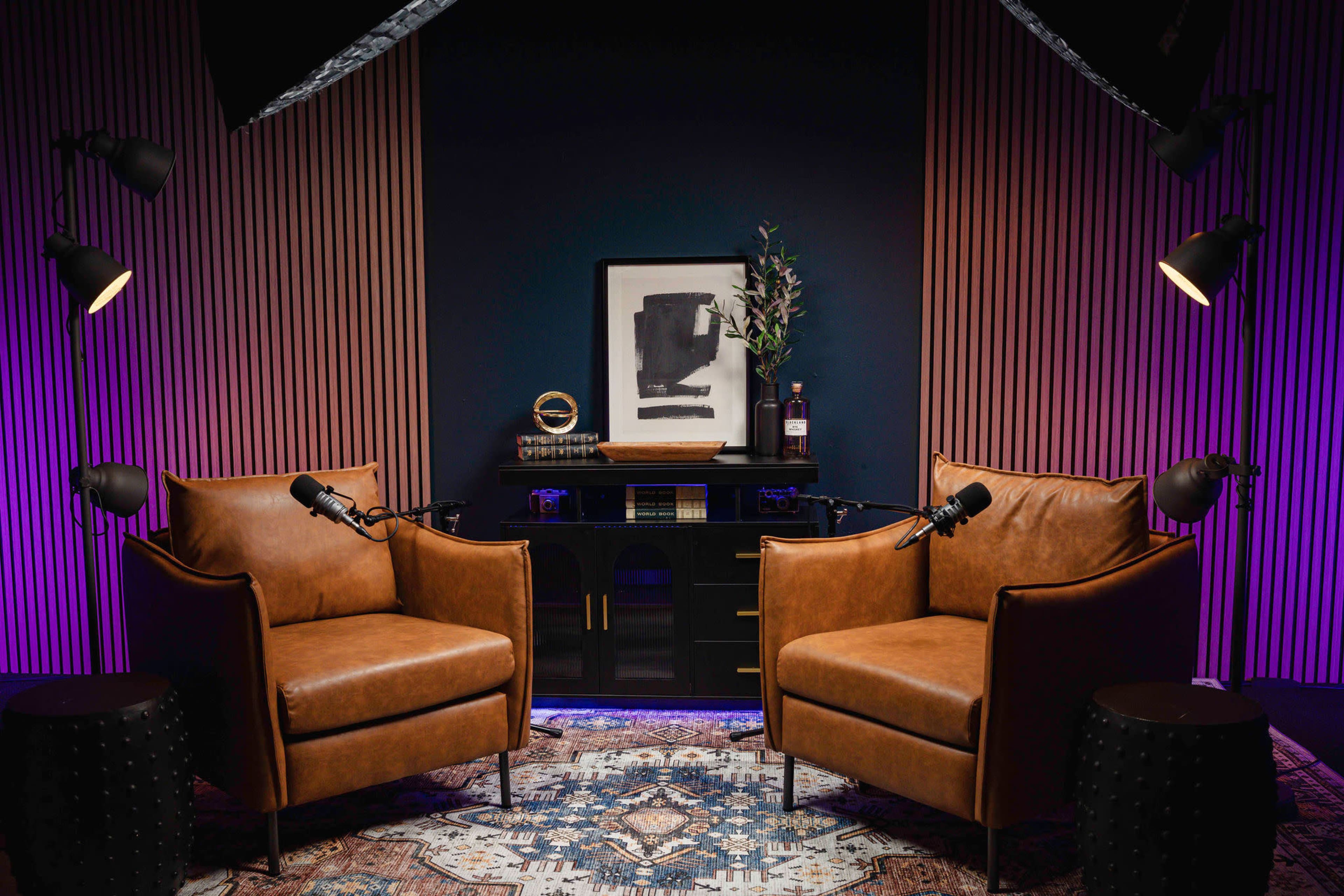 A modern recording studio setup features two leather chairs, a desk with decorative items, and two floor lamps, all set against a dark background with wooden paneling.