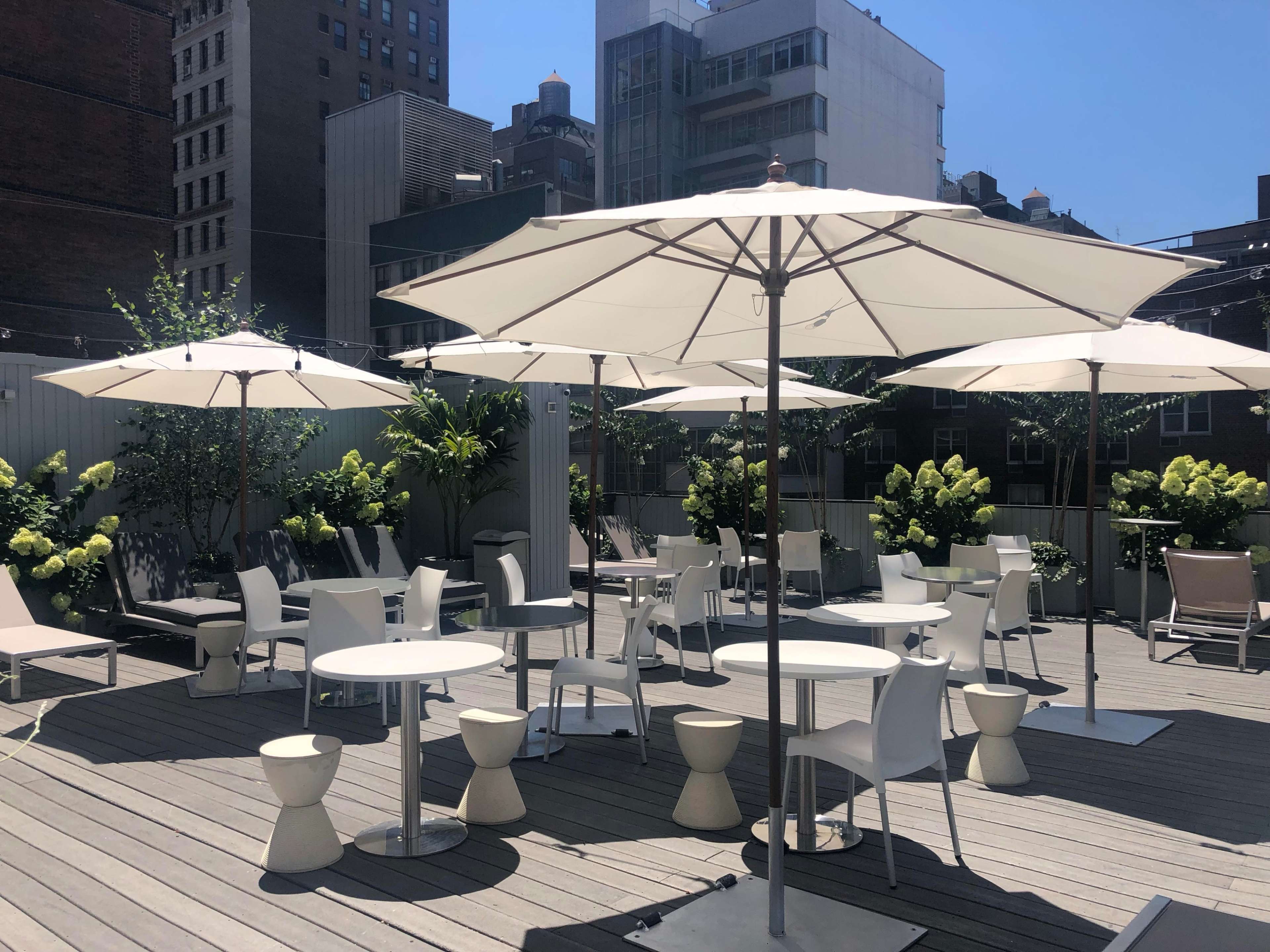 A rooftop space features white umbrellas, tables, and chairs surrounded by greenery and buildings under a clear blue sky.