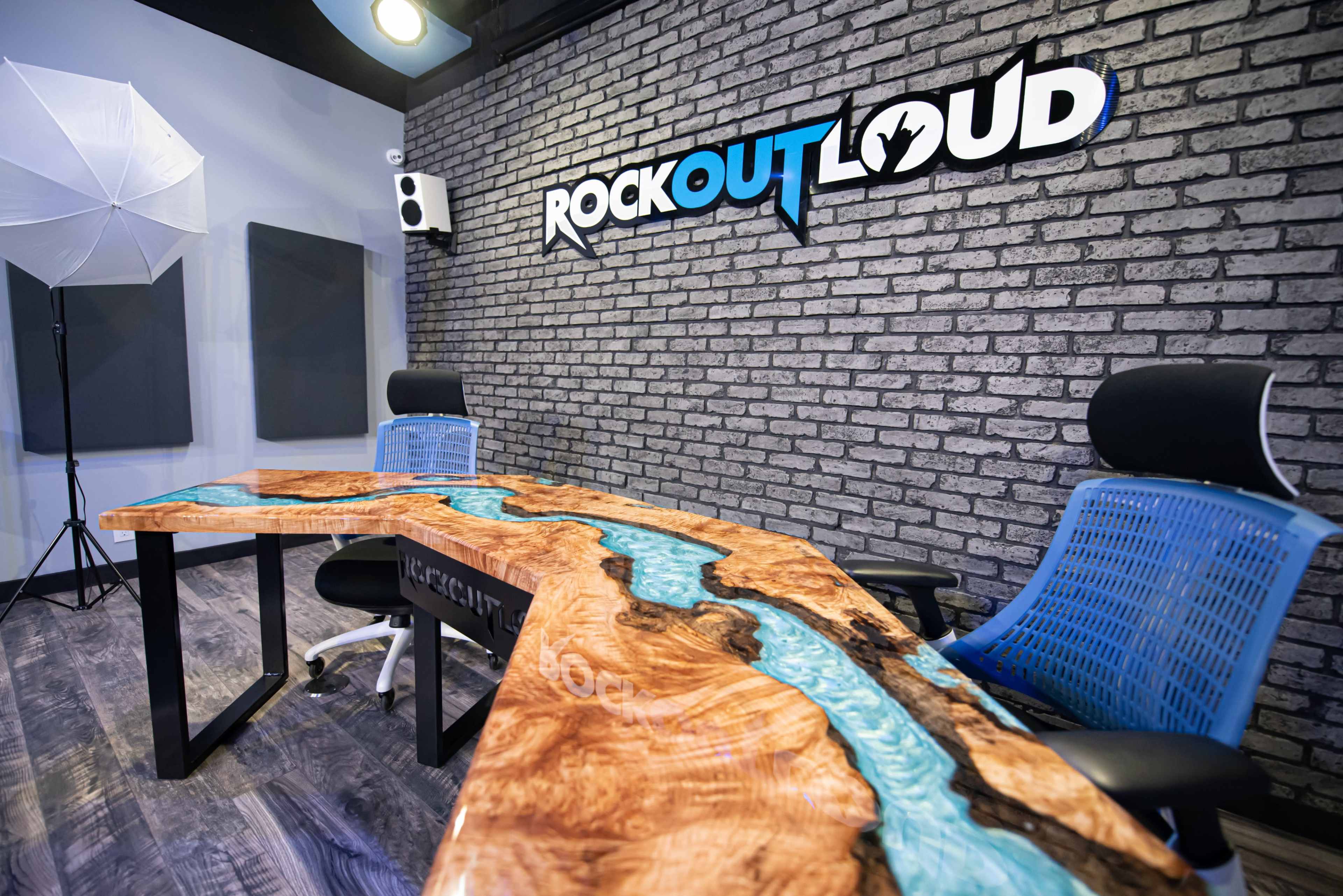 A modern workspace featuring a unique wooden conference table with a blue resin river design, accompanied by two ergonomic chairs and a wall with "ROCK OUT LOUD" signage.