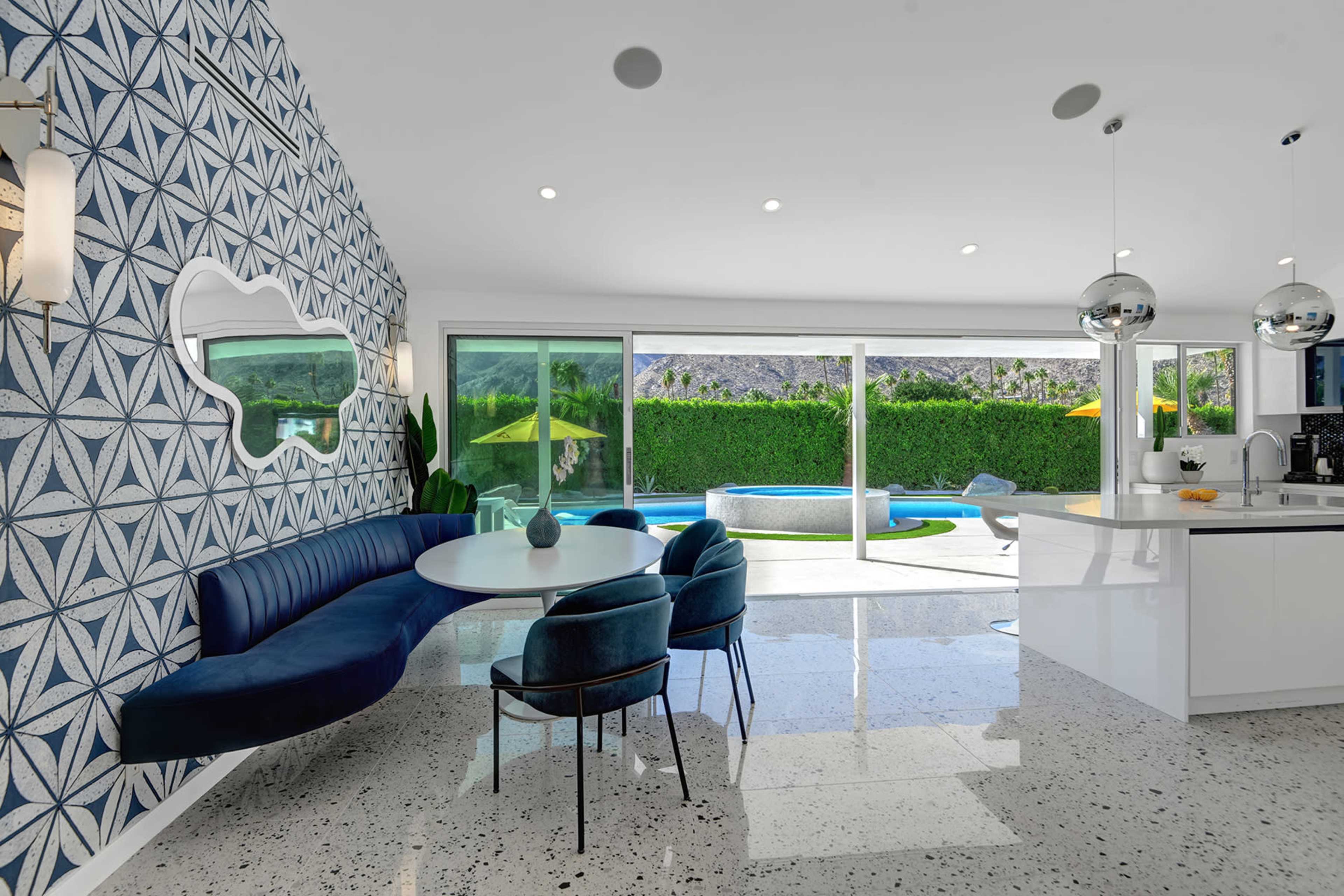 A modern kitchen and dining area feature a blue velvet banquette, a round table, and large sliding glass doors leading to a pool and patio outside.