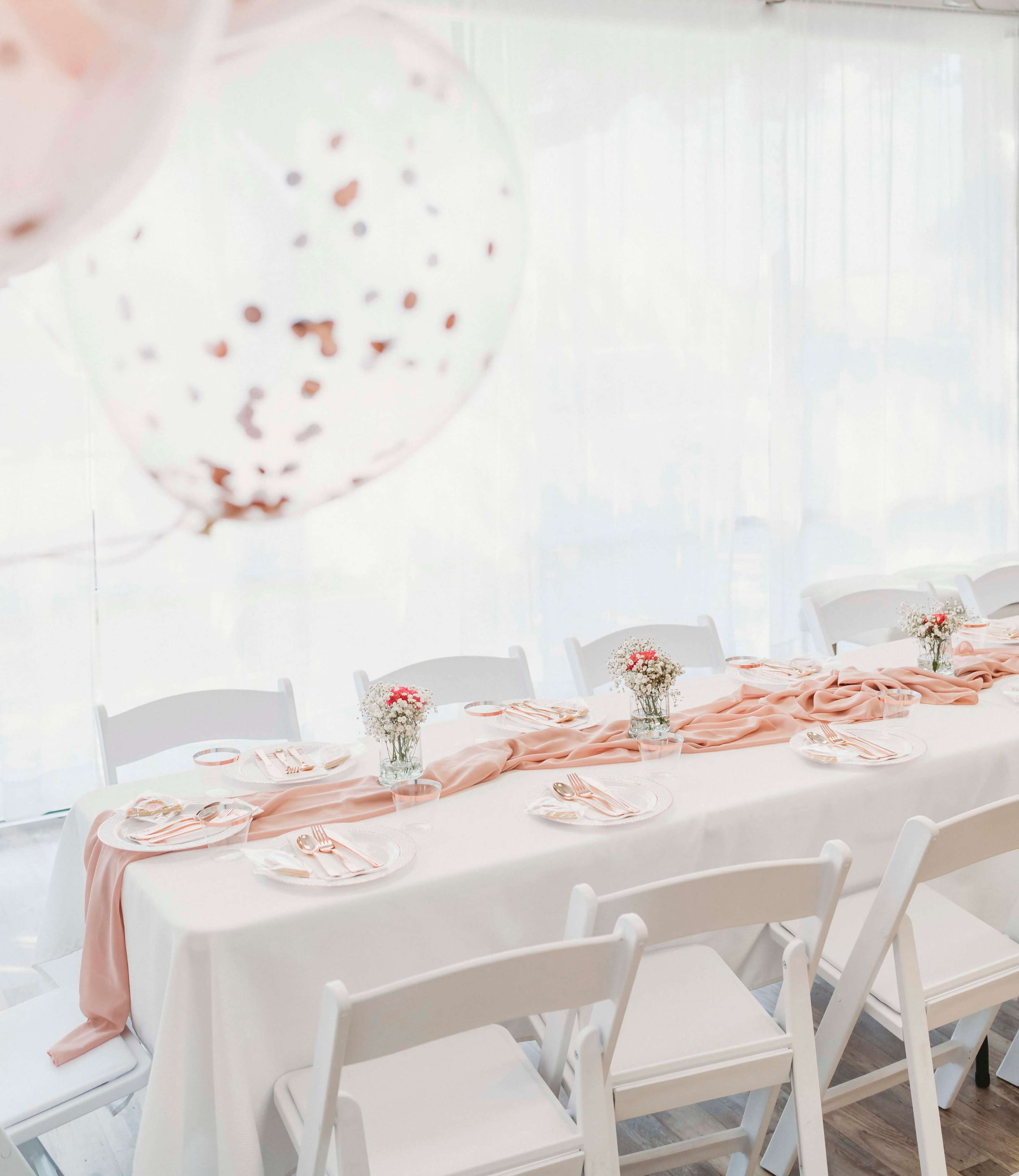 A long table is elegantly set with white tablecloths, light pink runners, and small floral centerpieces, surrounded by white chairs.