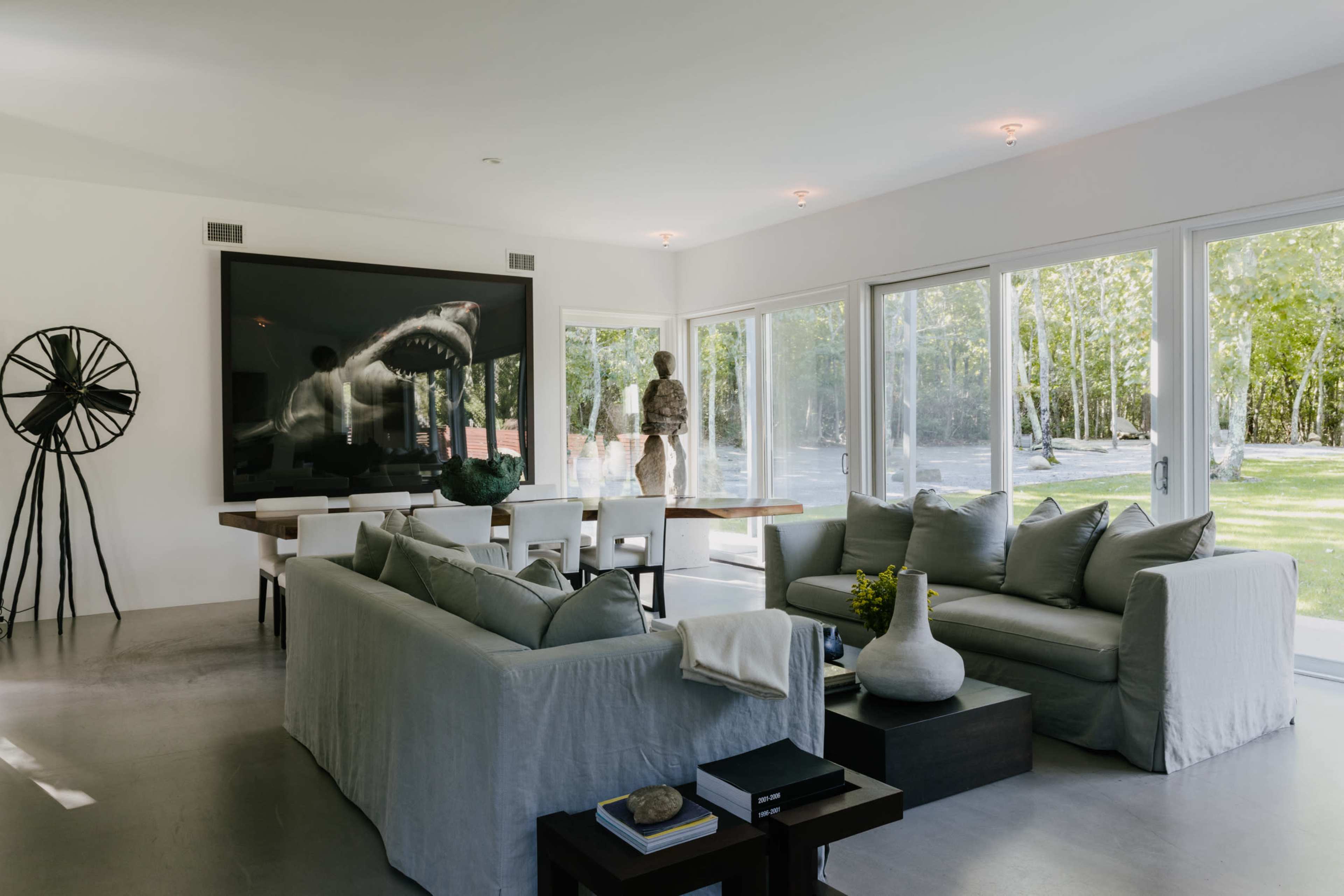 A modern living room featuring a large shark artwork, a dining table, and large glass windows overlooking a green outdoor area.