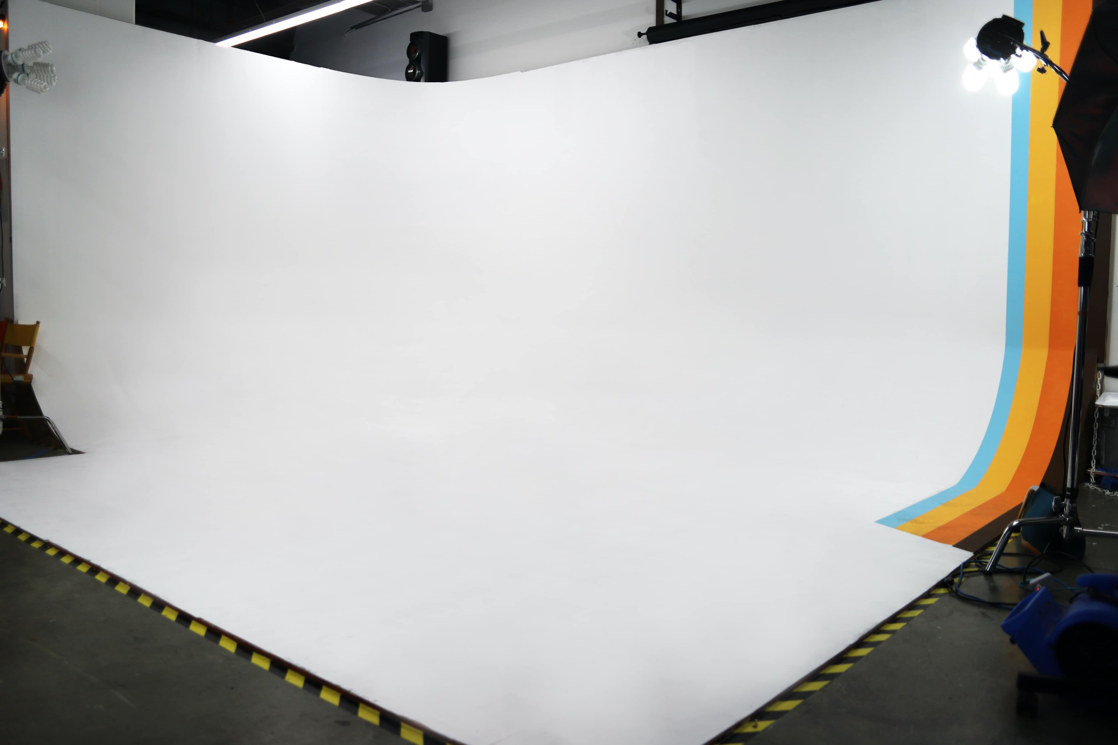 The image shows a photography studio with a curved white backdrop featuring orange and blue stripes, along with studio lighting equipment.