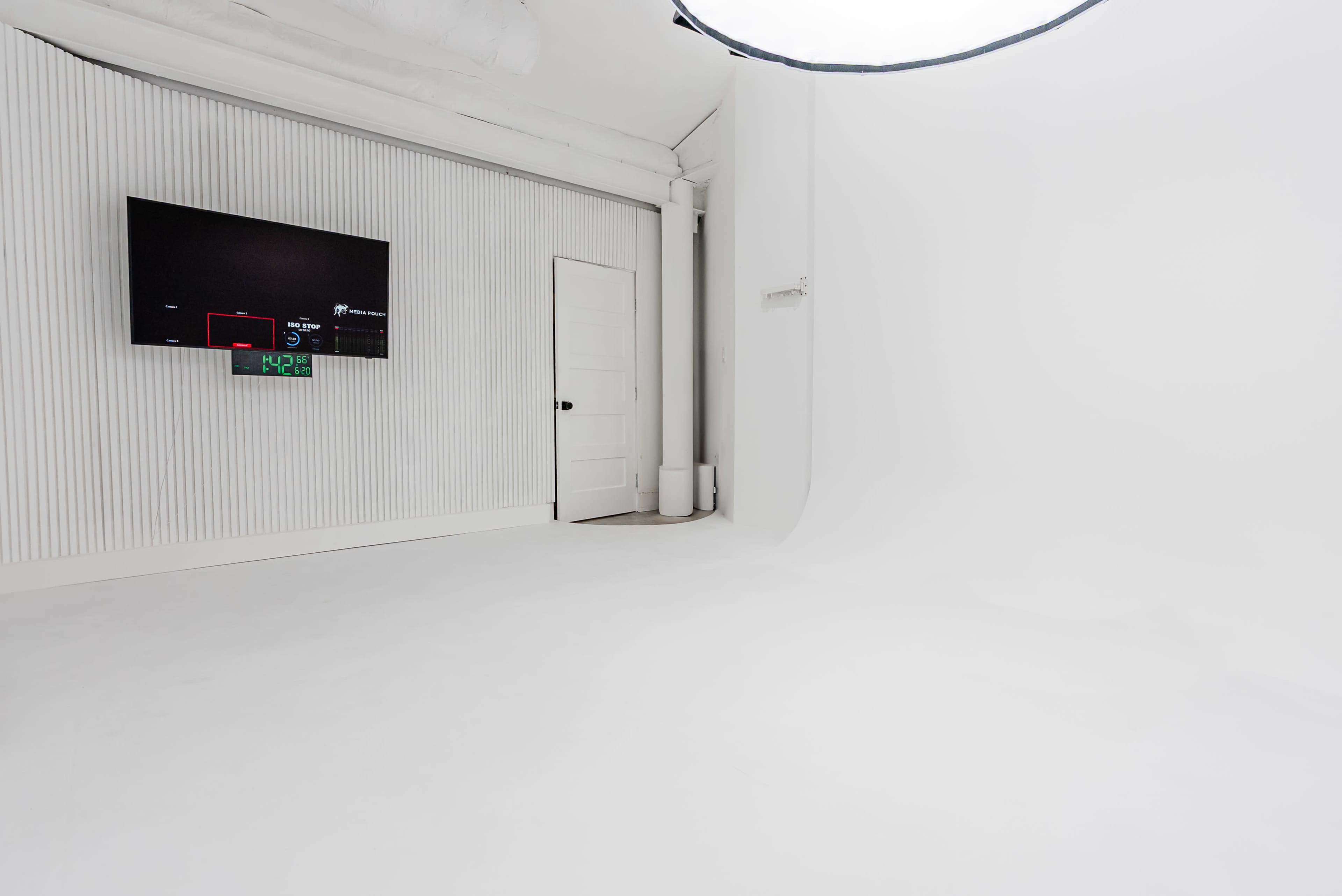 The image shows a spacious, minimalistic studio with a white floor, white walls, and a large screen mounted on one wall.