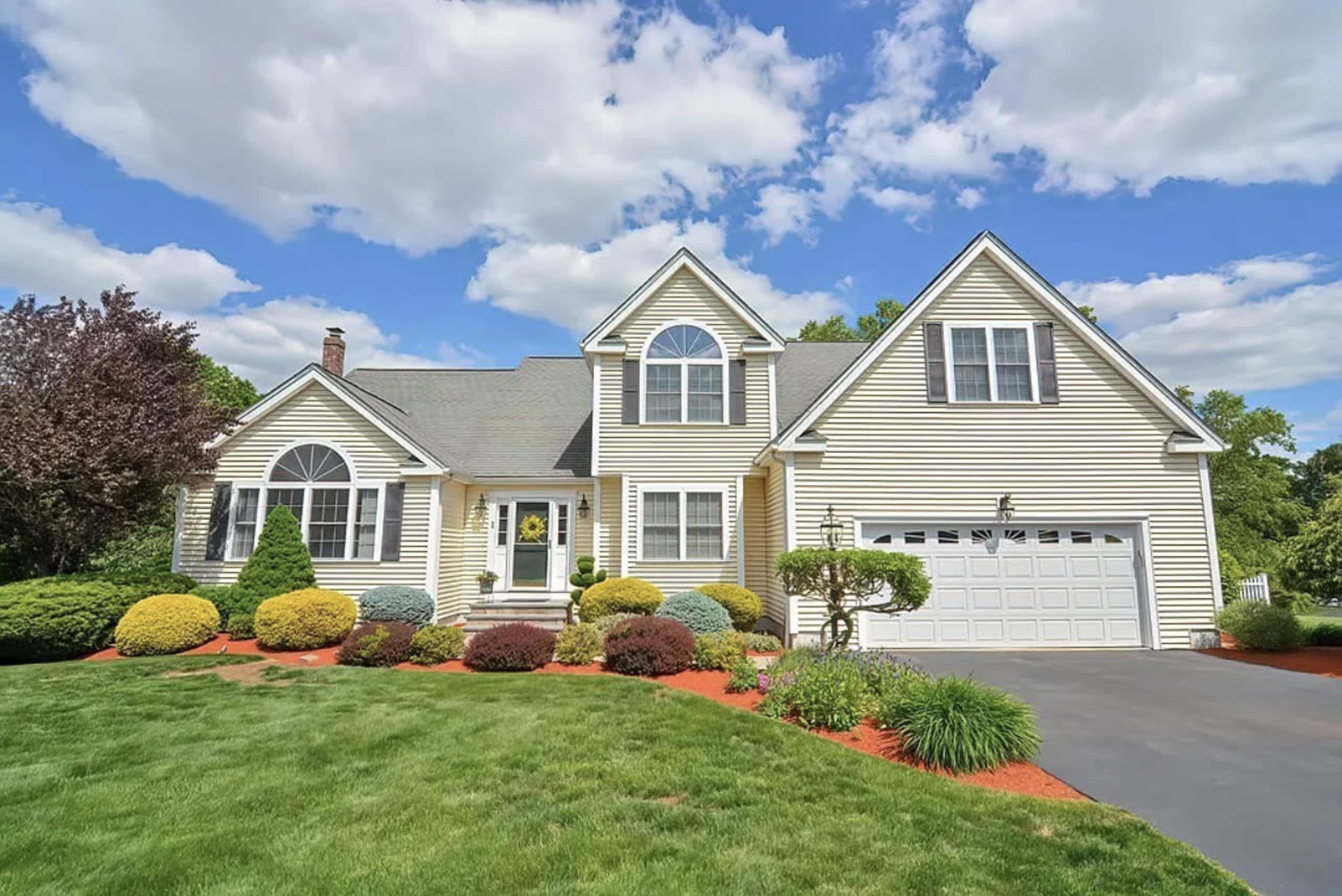 A two-story, yellow-sided house with a landscaped front yard and a driveway leading to a garage.