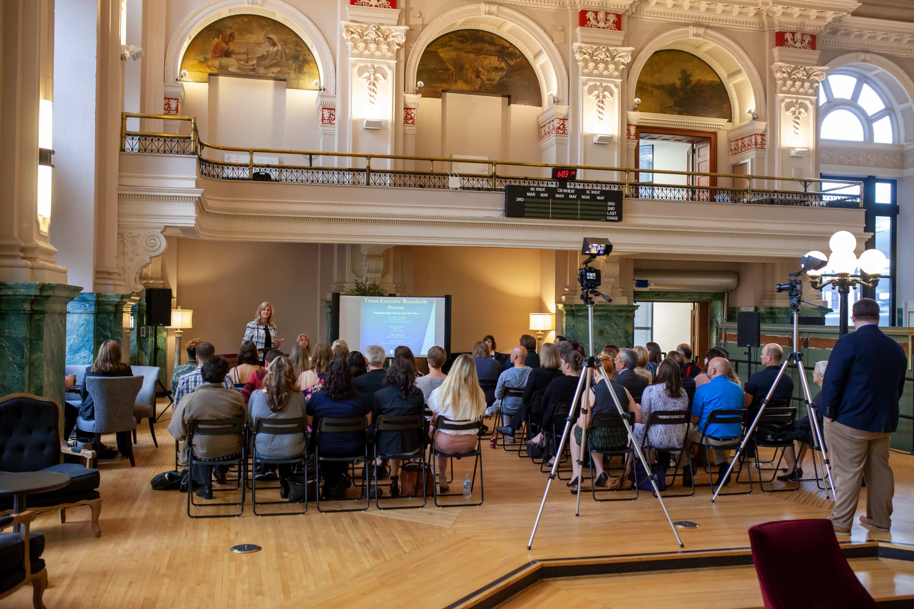 A group of individuals is seated in a hall, listening to a speaker in front of a presentation screen, with large windows and decorative architecture in the background.