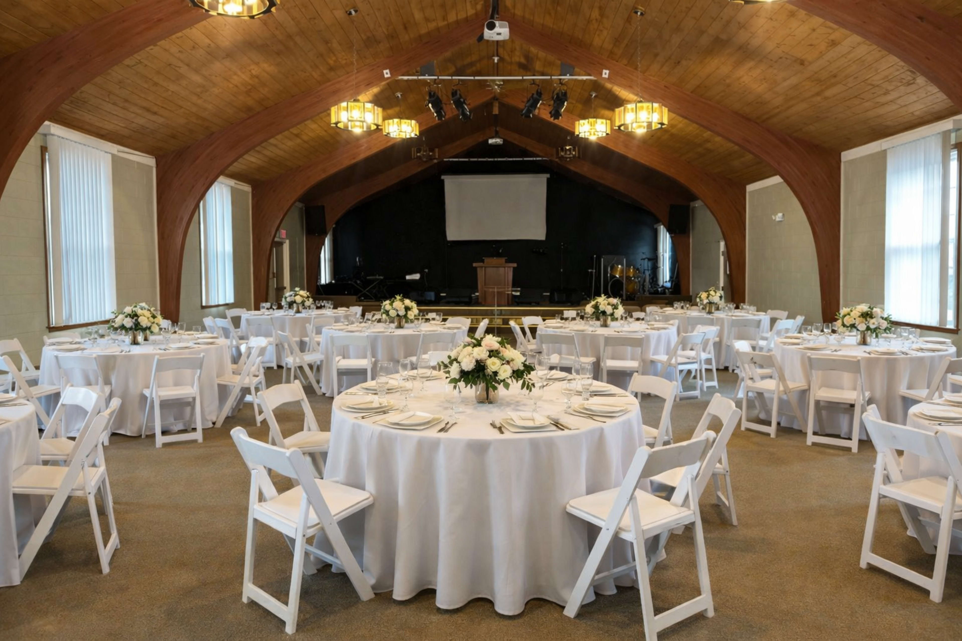 The image shows a spacious banquet hall set with round tables covered in white tablecloths, each adorned with floral centerpieces, and equipped with a stage at the front.