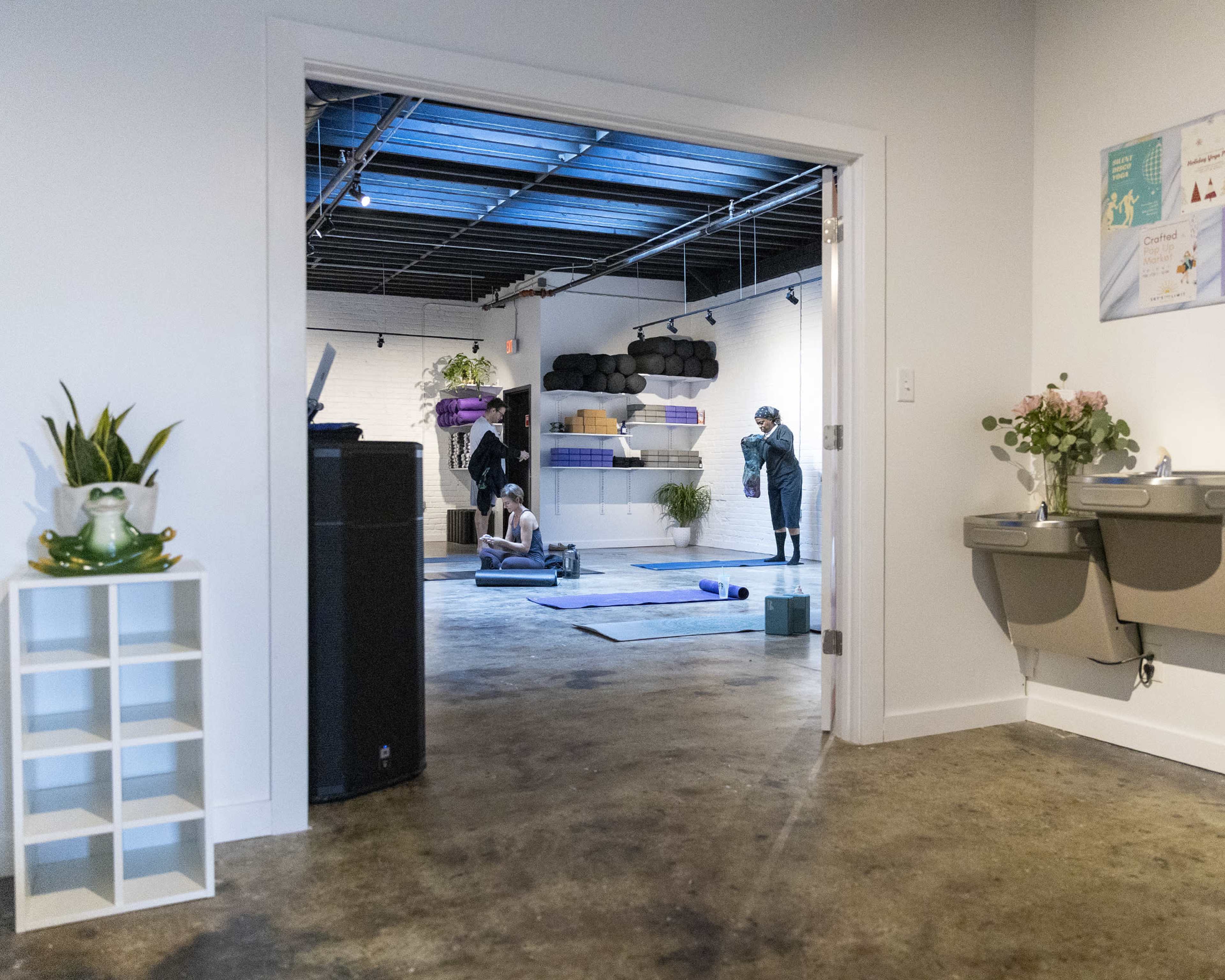 A brightly lit fitness studio features individuals practicing yoga on mats, with workout equipment and plants visible in the background.