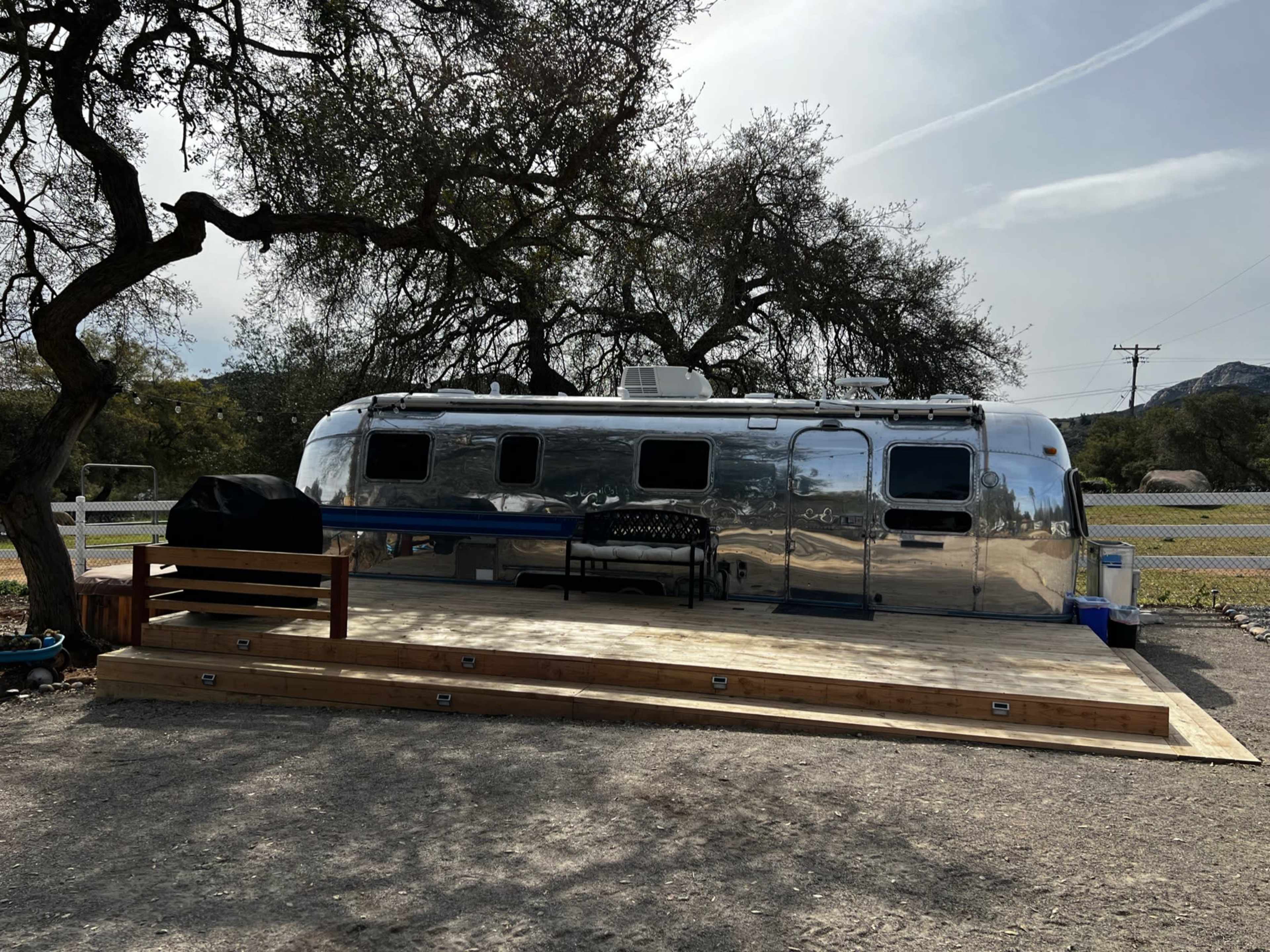 A polished Airstream trailer is parked beside a wooden deck under the shade of an oak tree.
