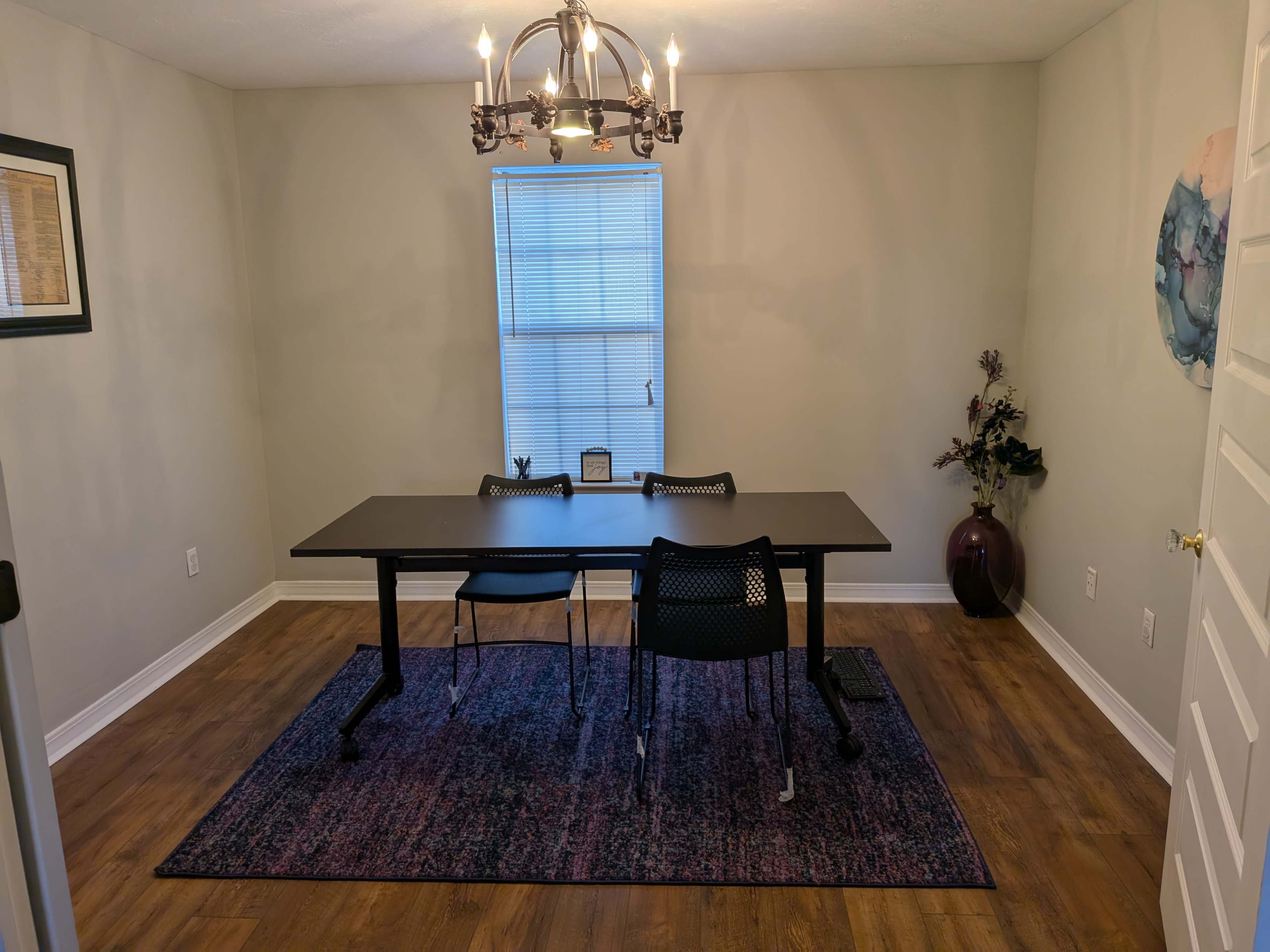 A simple room features a rectangular table with two black chairs, a window allowing light to enter, and a decorative plant in the corner.