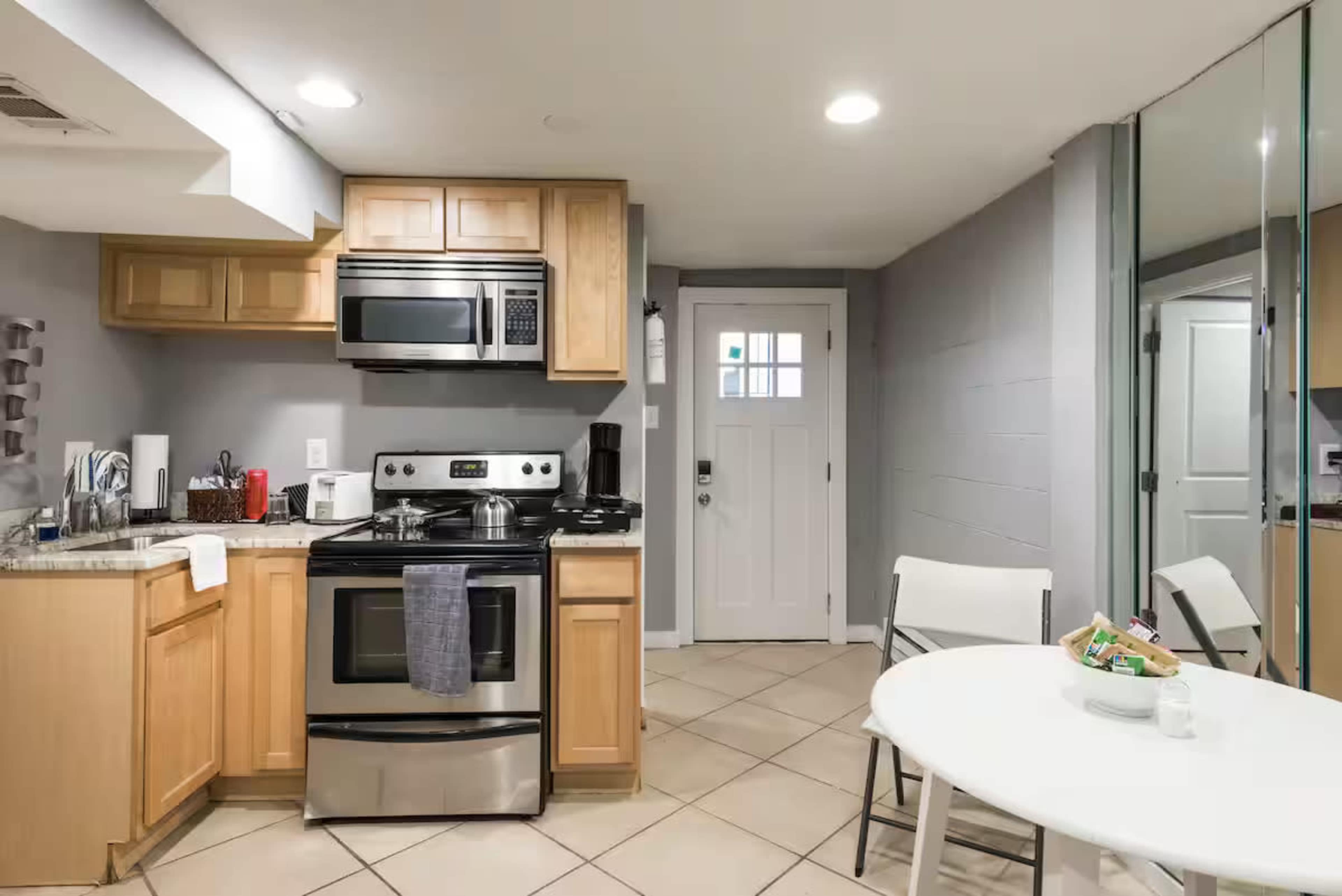 The image shows a compact kitchen with wooden cabinets, stainless steel appliances, and a small dining area featuring a round table and chairs.