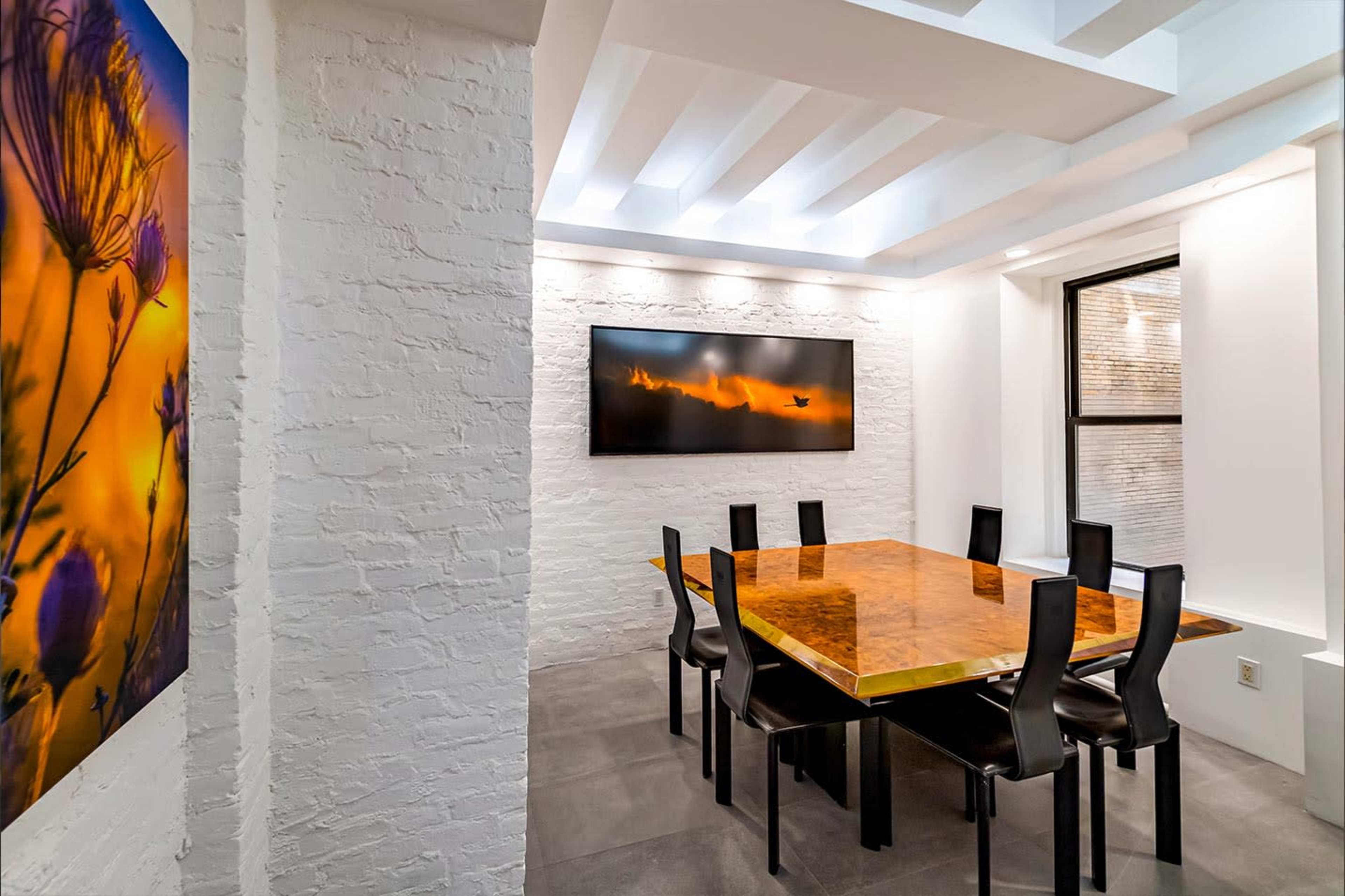 The image shows a modern dining room featuring a large wooden table surrounded by black chairs, white brick walls, and a wall-mounted television.