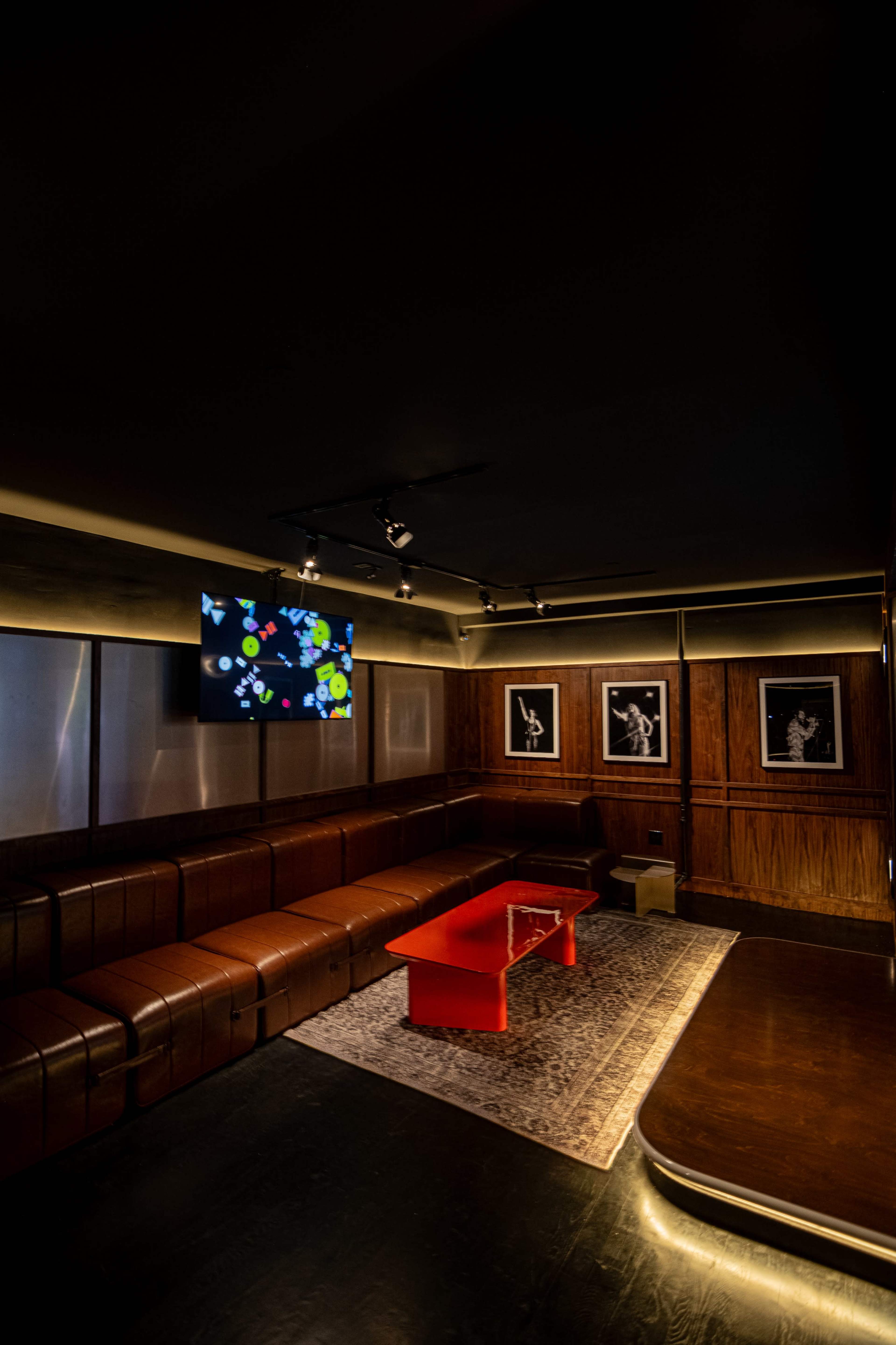 The image shows a dimly lit lounge area with a red coffee table, leather seating, a television displaying colorful graphics, and framed black-and-white art on the walls.