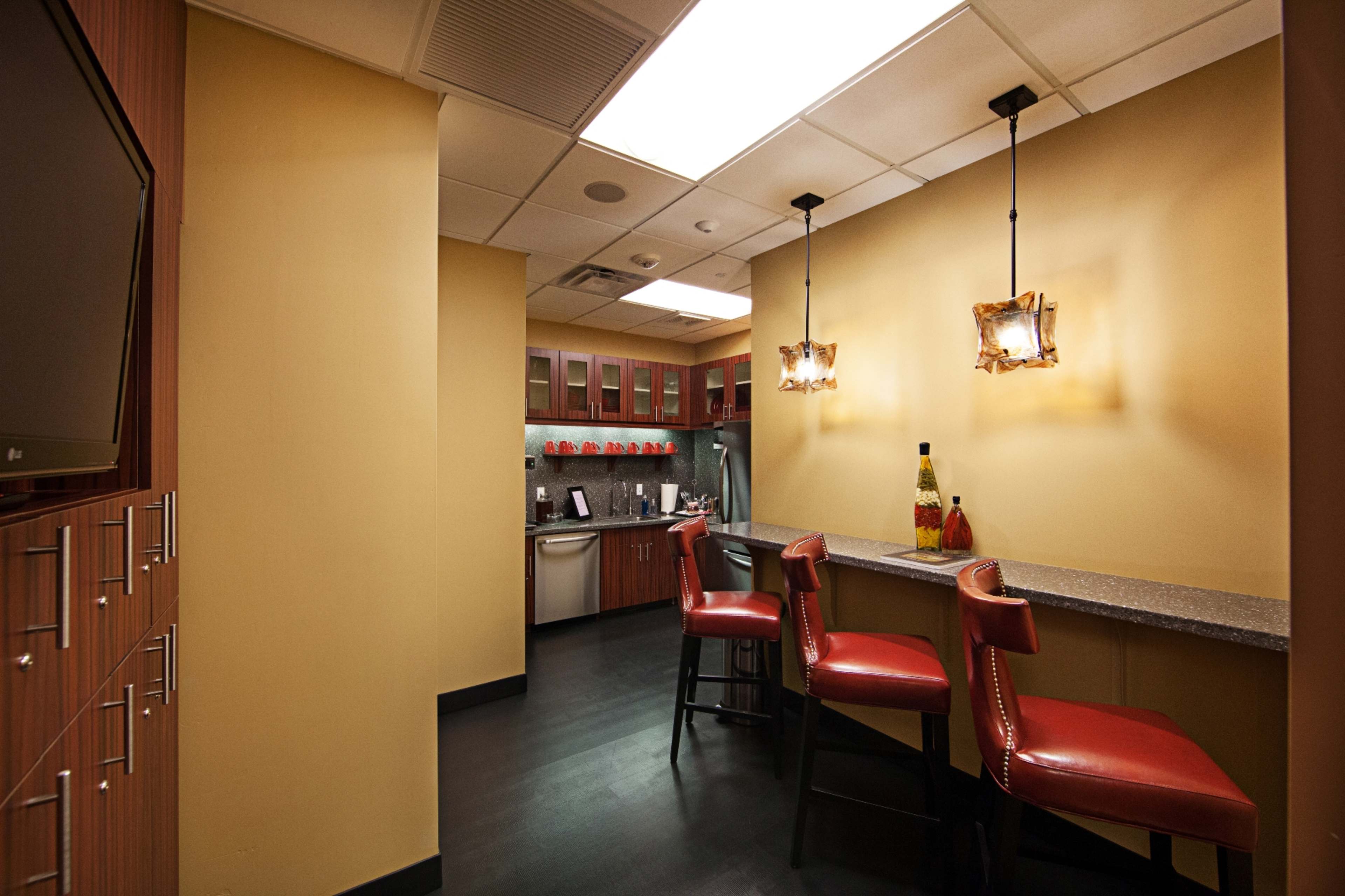 A modern kitchen area with a bar counter and three red stools, featuring cabinets and a small appliance area in a well-lit space.