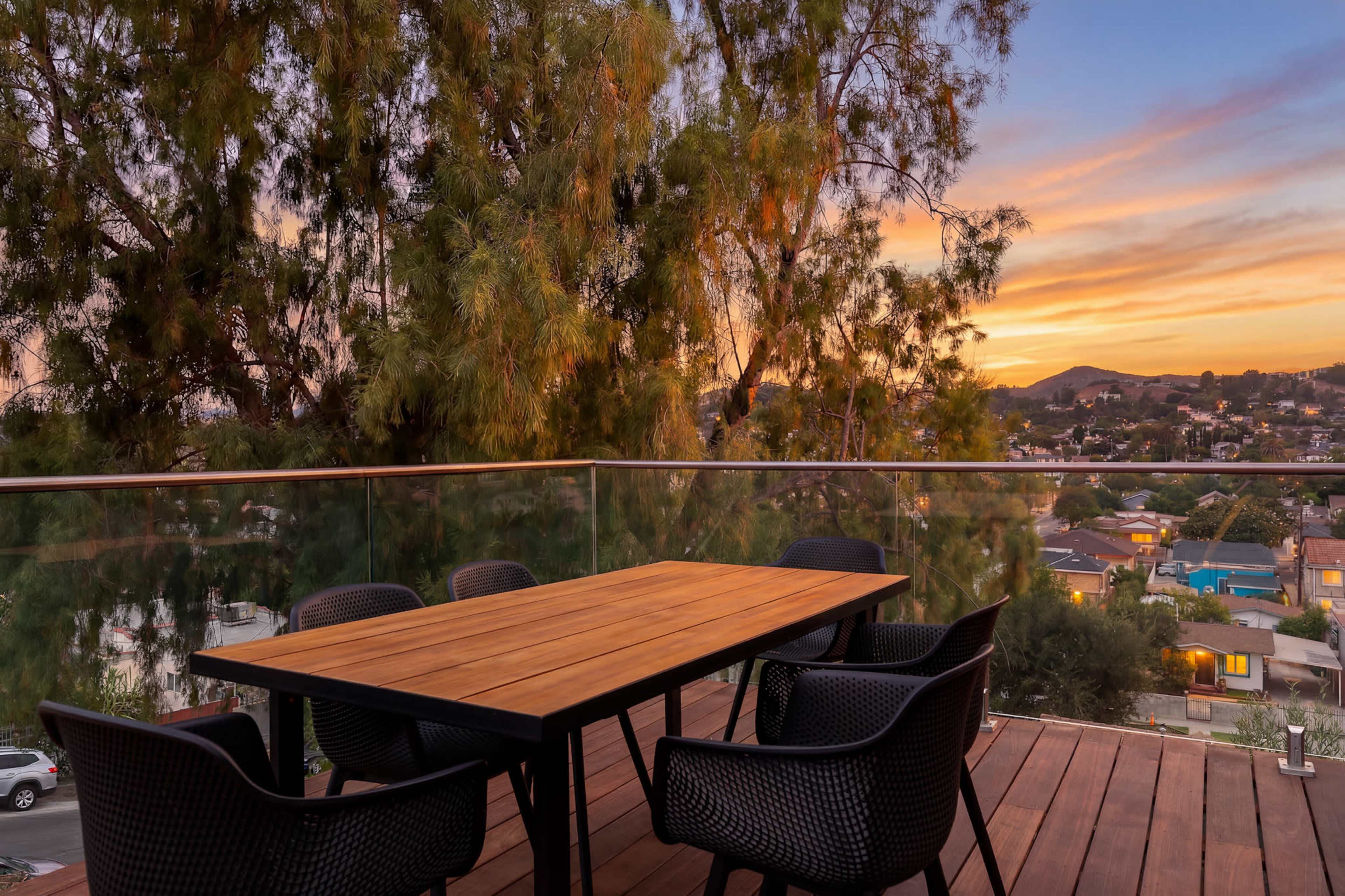 A wooden table surrounded by black chairs sits on a balcony with a view of a sunset over a neighborhood.