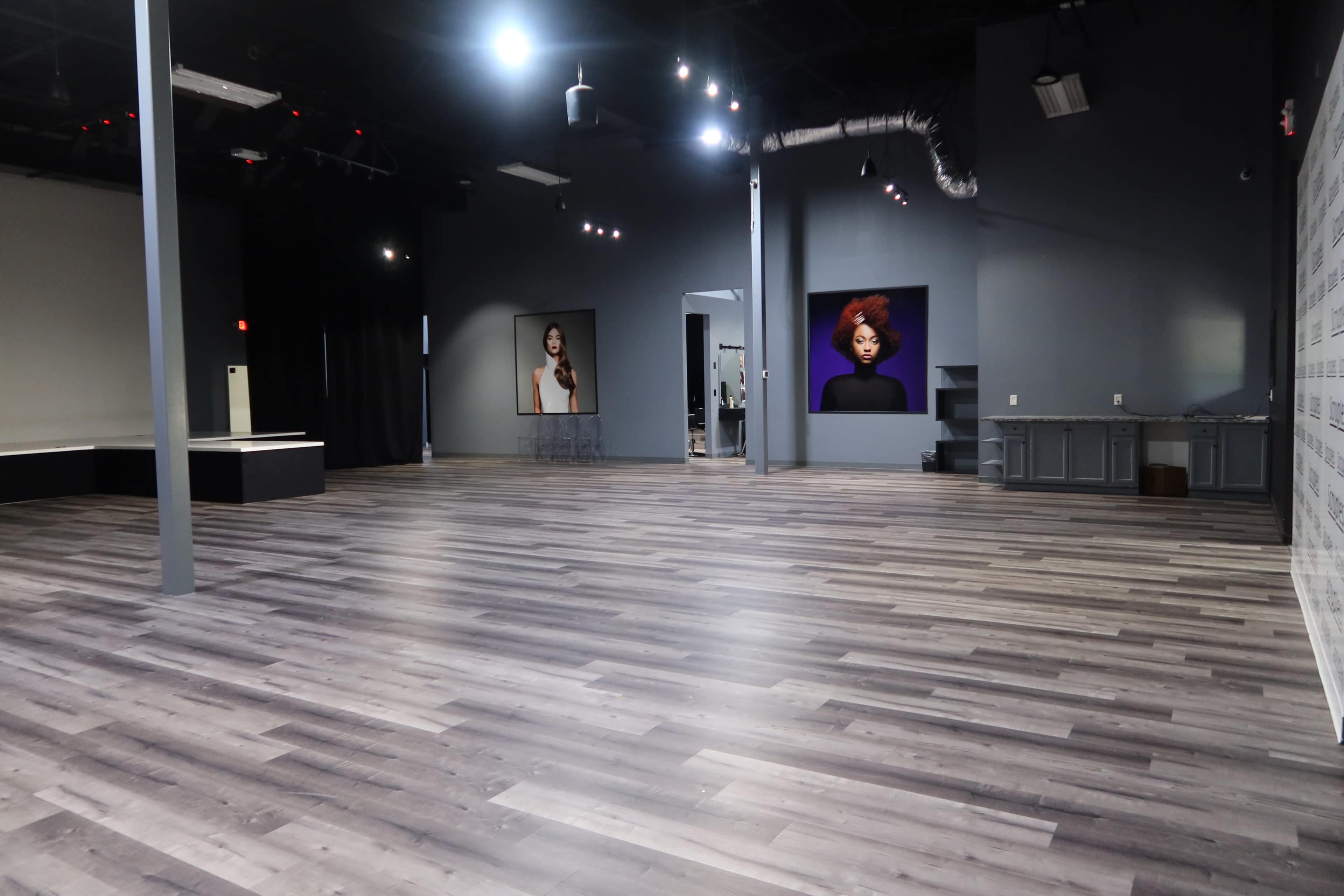 The image shows a spacious, well-lit studio interior with wooden flooring, a gray wall featuring two large portraits, and a counter along one side.
