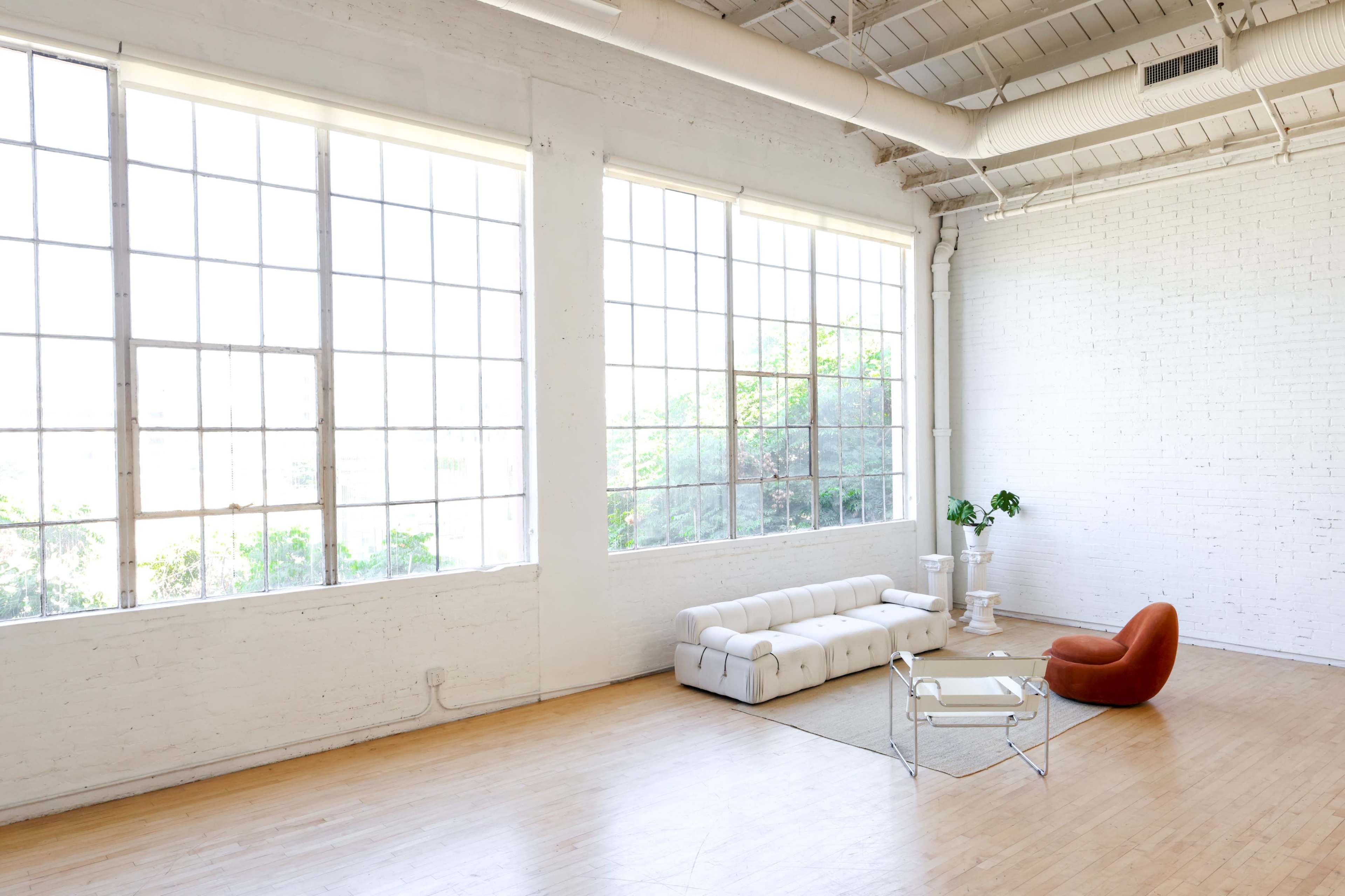 The image shows a spacious, brightly lit room with large windows, a white couch, an orange chair, and a small table.