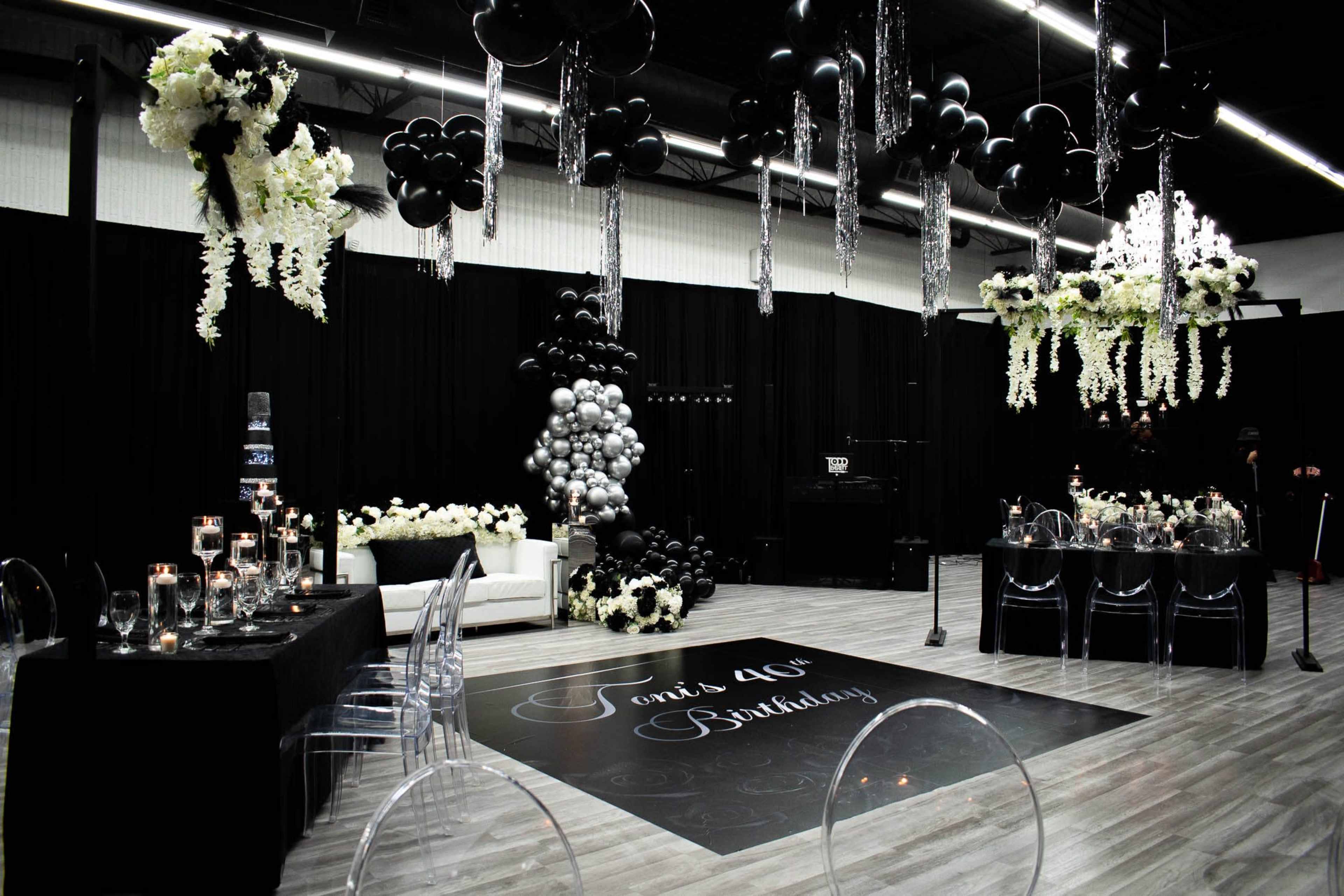 The scene displays a decorated venue for a birthday party, featuring black and white balloons, floral arrangements, a dining table set with elegant tableware, and a central dance floor with the text "Toni's 40th Birthday."