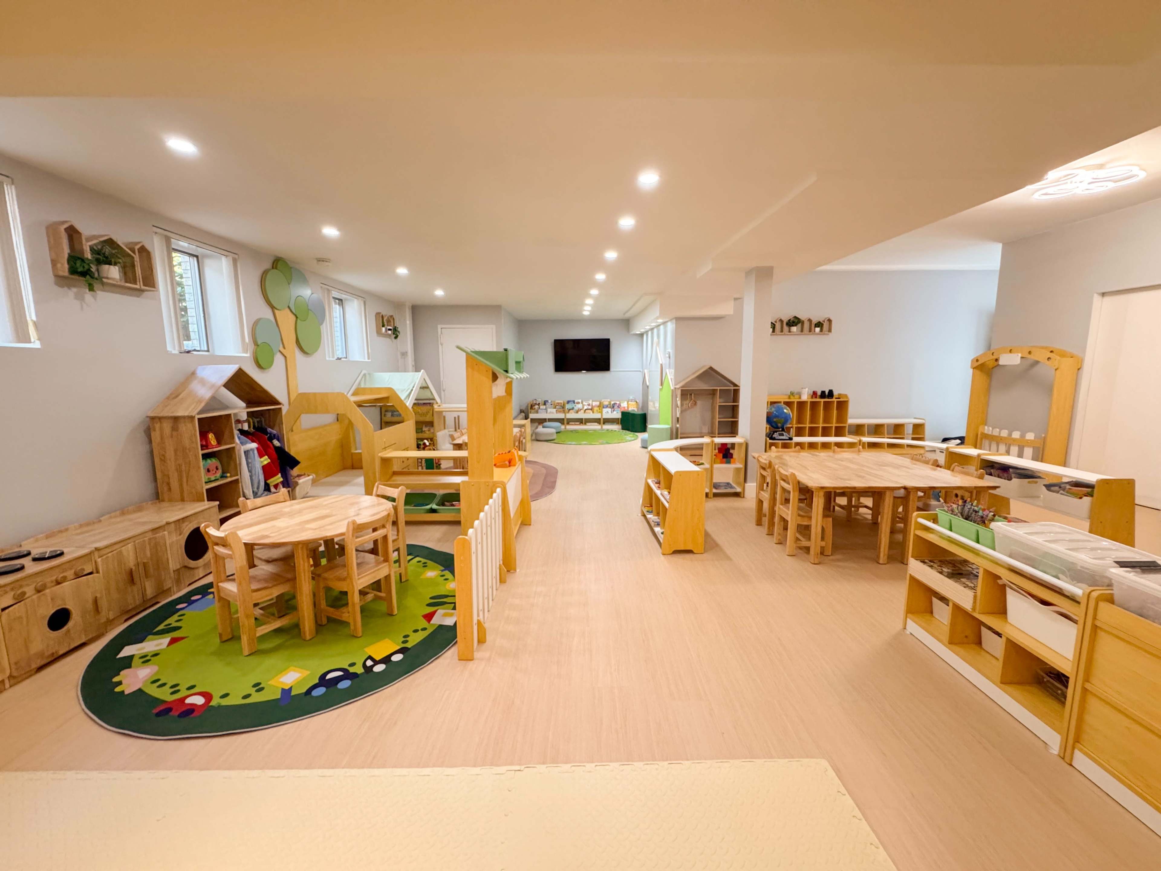 The image shows a brightly lit early childhood classroom with wooden tables, chairs, and various play structures arranged around the room.