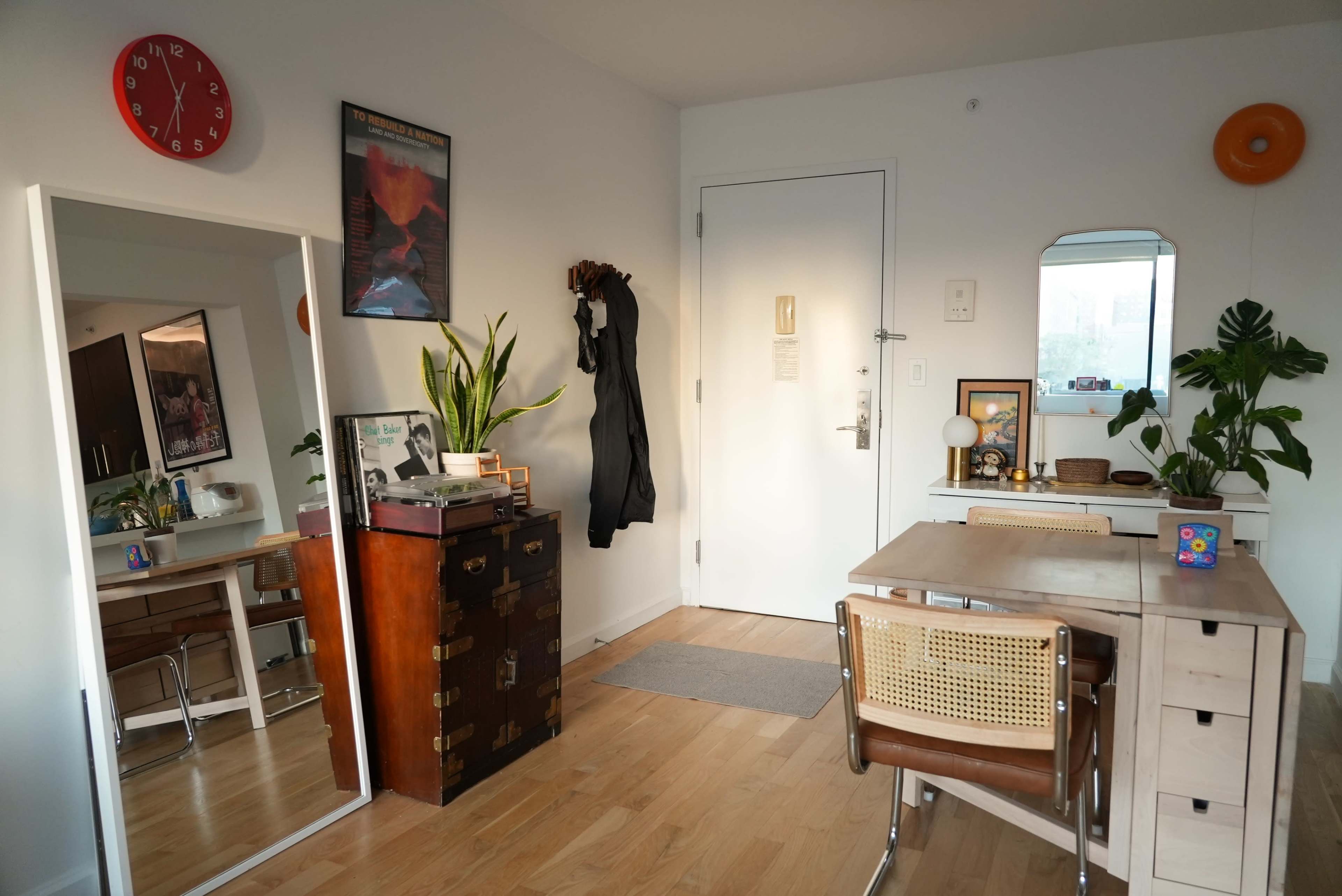 The image shows a well-lit room featuring a mirror, a potted plant, a table with chairs, and a door with a simple decorative arrangement.