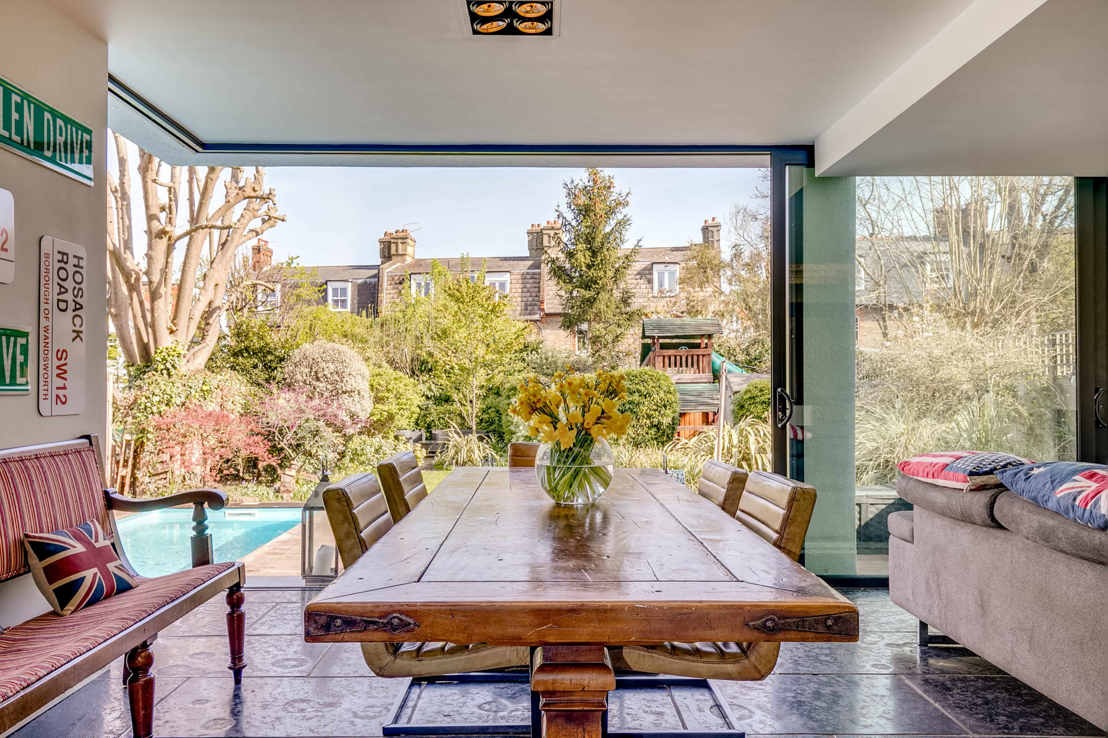 A wooden dining table with a vase of yellow flowers sits in a well-lit room that opens to a garden featuring a pool and various greenery.