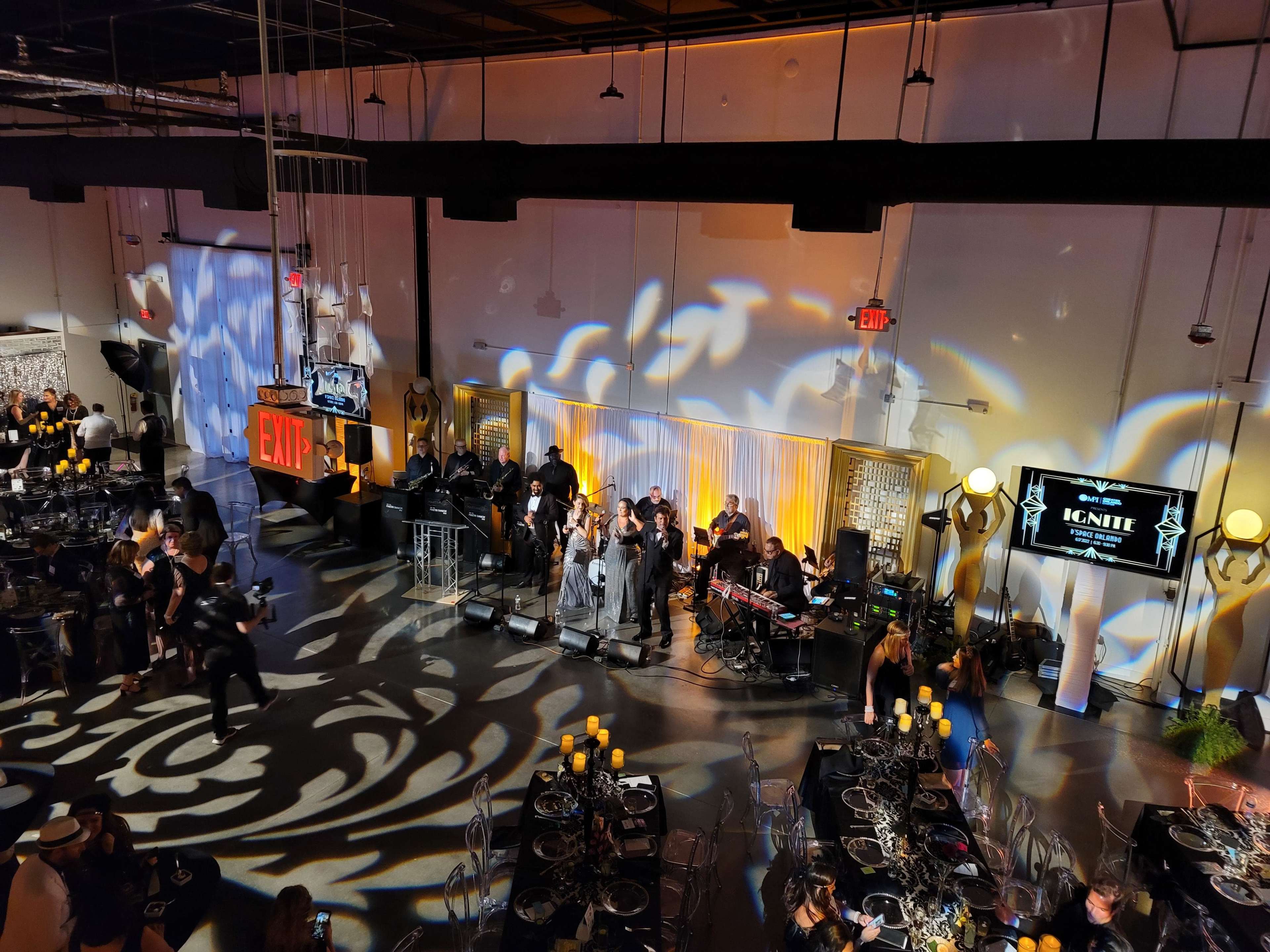 A large event space is set up with tables and chairs, featuring a stage where a band is preparing to perform, illuminated by dramatic lighting patterns on the floor.