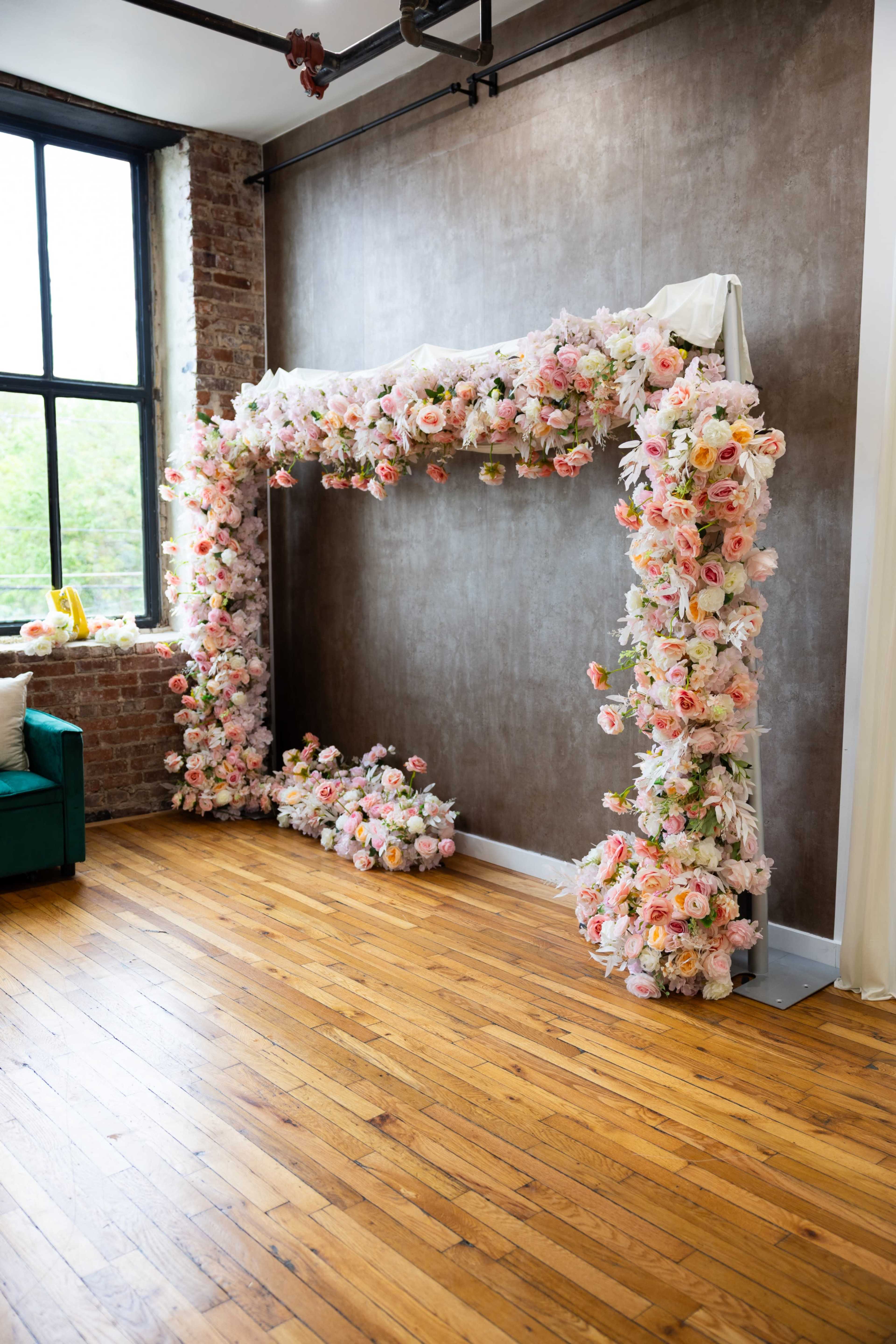 The image shows a floral arch adorned with pink and white flowers in an industrial-style room with large windows and wooden flooring.