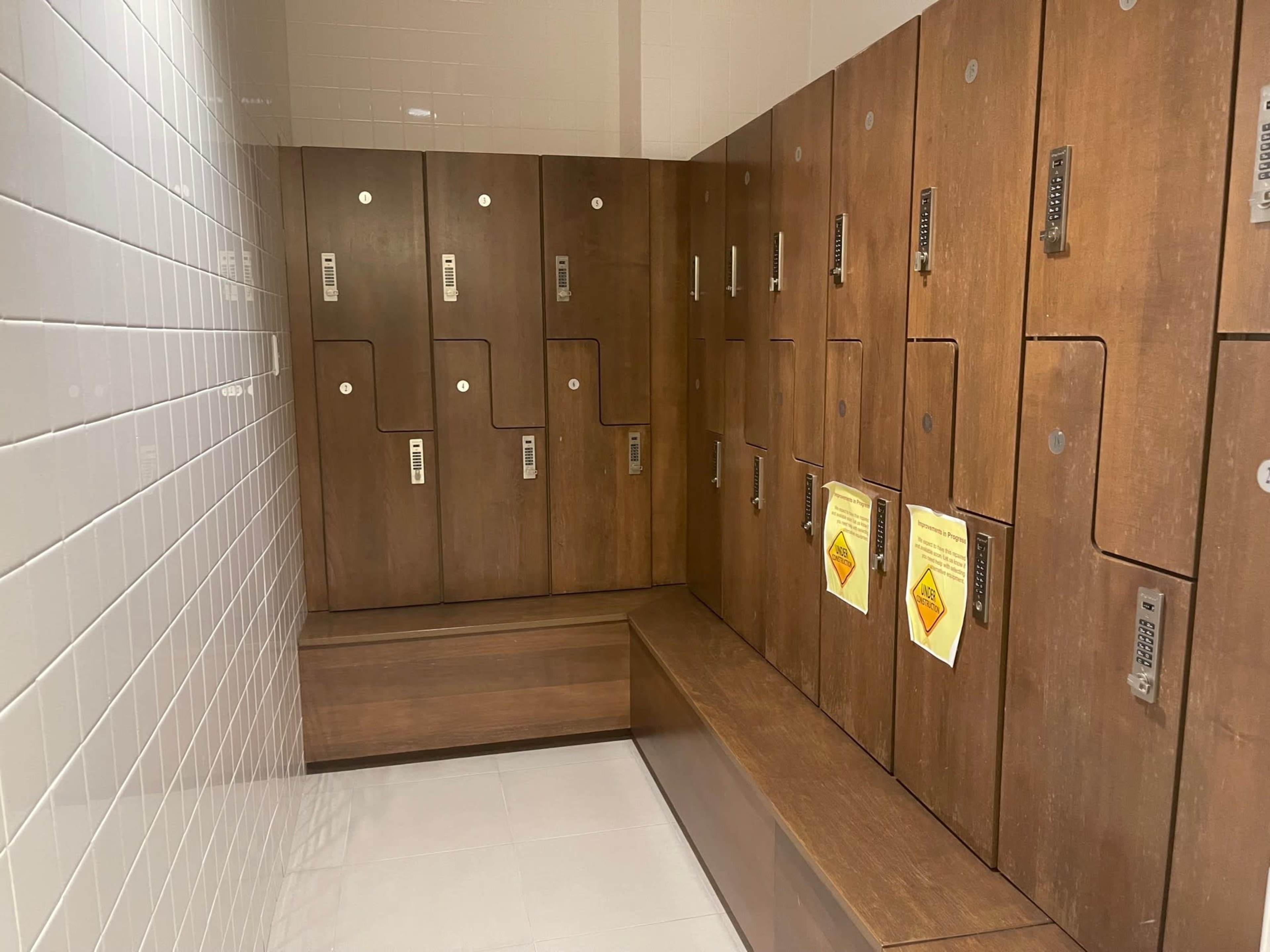 The image shows a row of wooden lockers with a bench in a locker room, featuring white tiled walls.