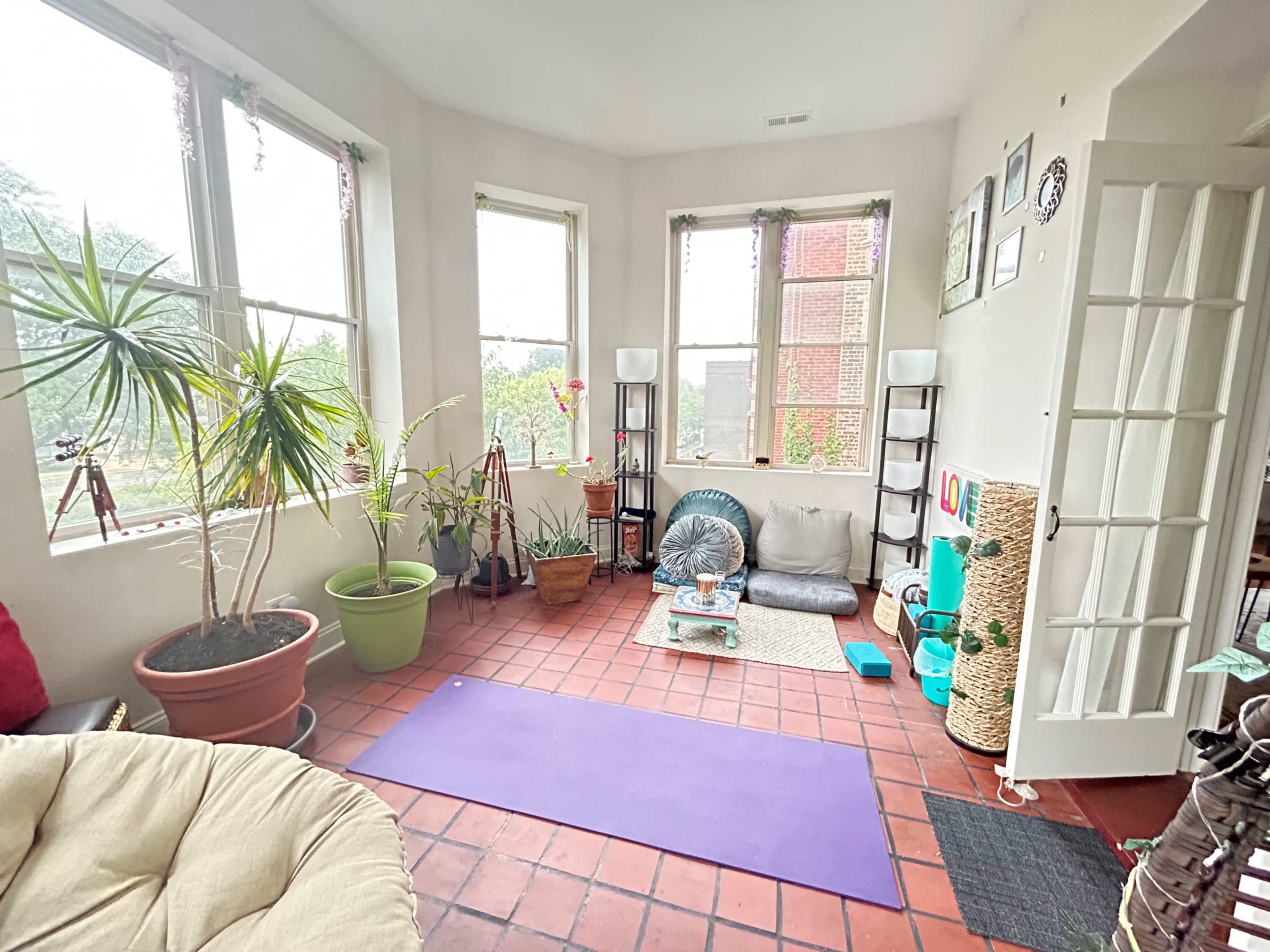 The image shows a bright sunroom with large windows, plants, a yoga mat, and various seating areas arranged around a tiled floor.