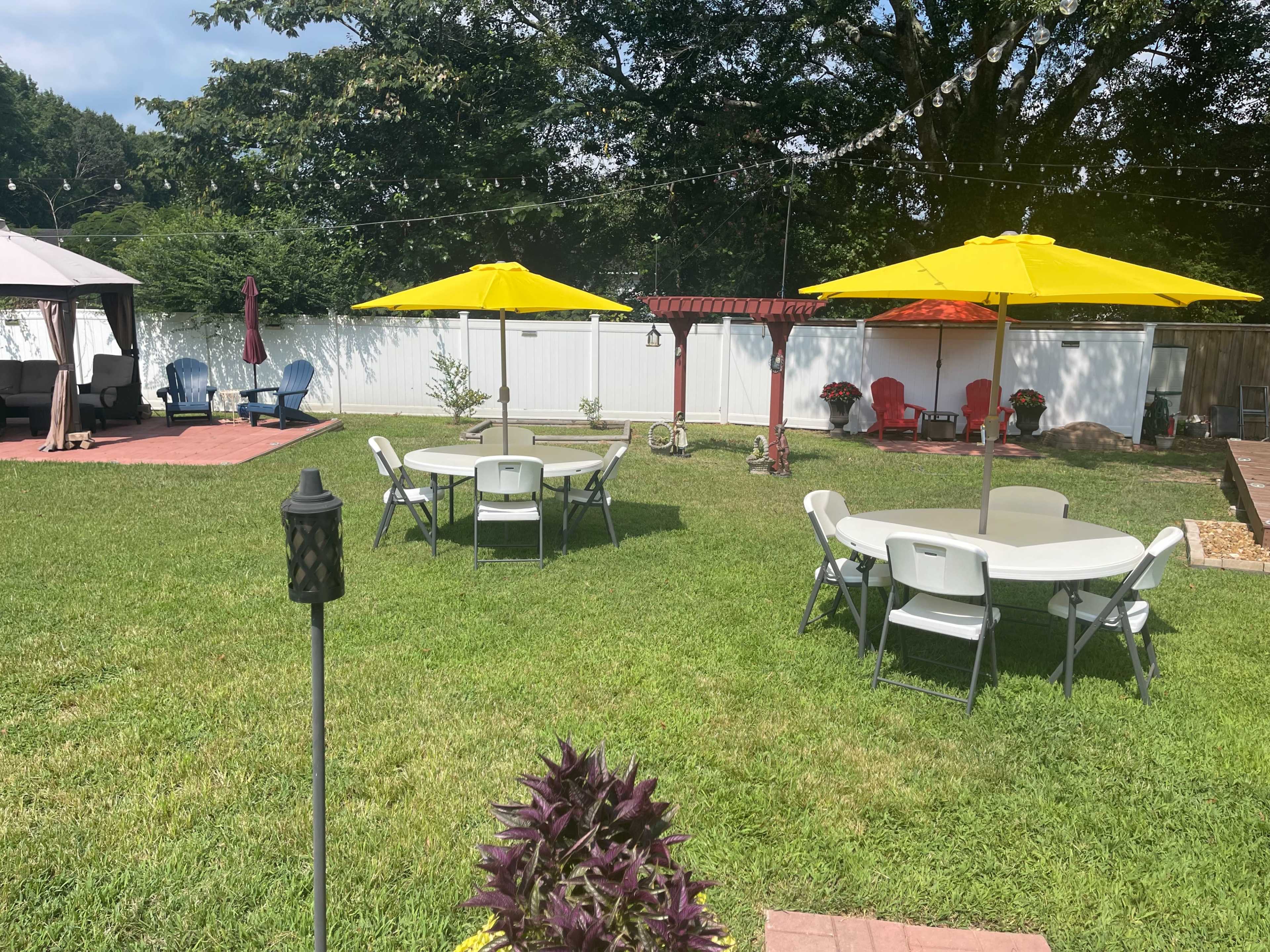 The image shows a backyard with several round tables under yellow umbrellas, surrounded by green grass and various seating arrangements.