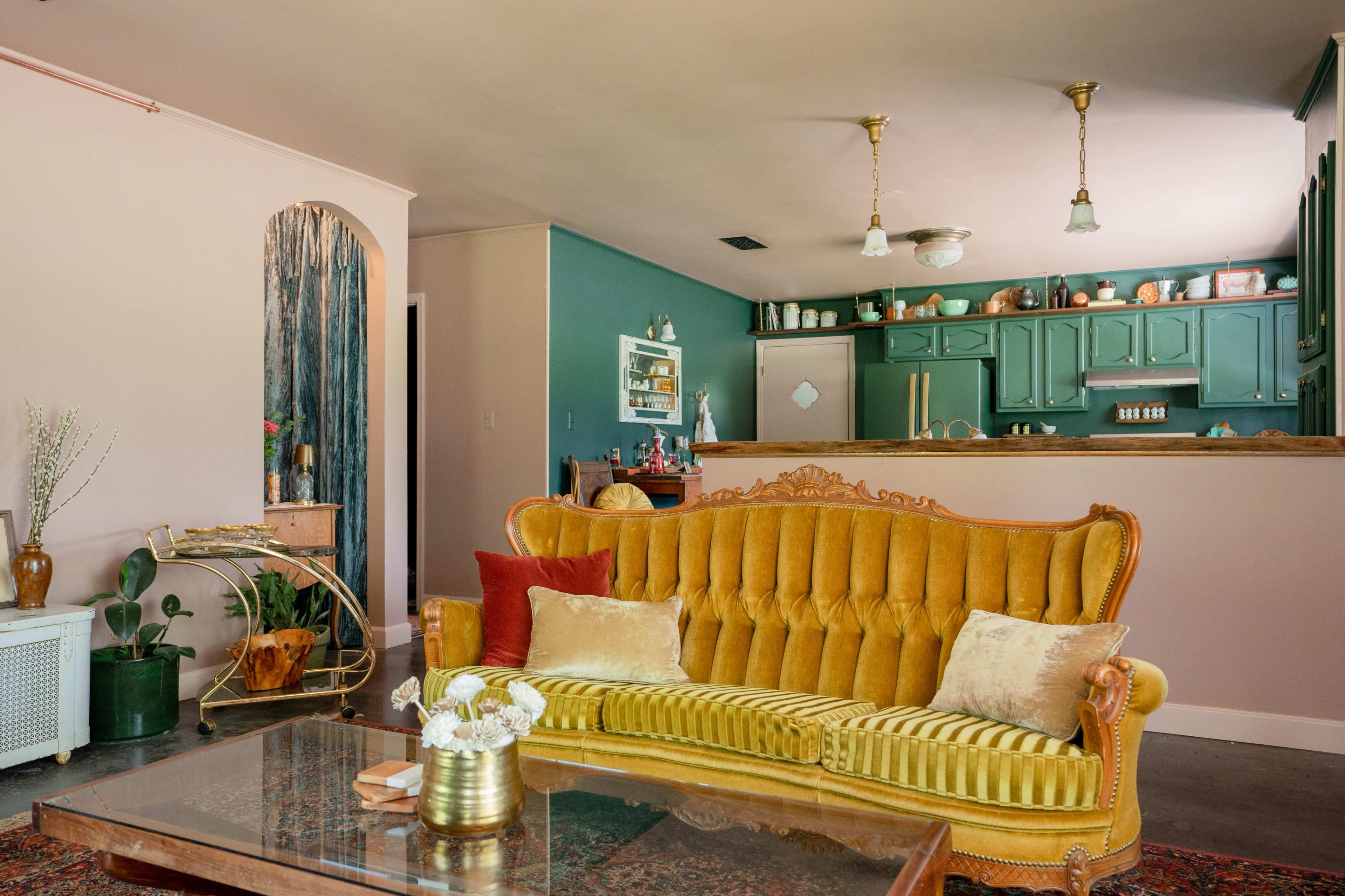 A vintage yellow sofa is centered in a living room with teal walls, a glass coffee table, and a kitchen visible in the background.