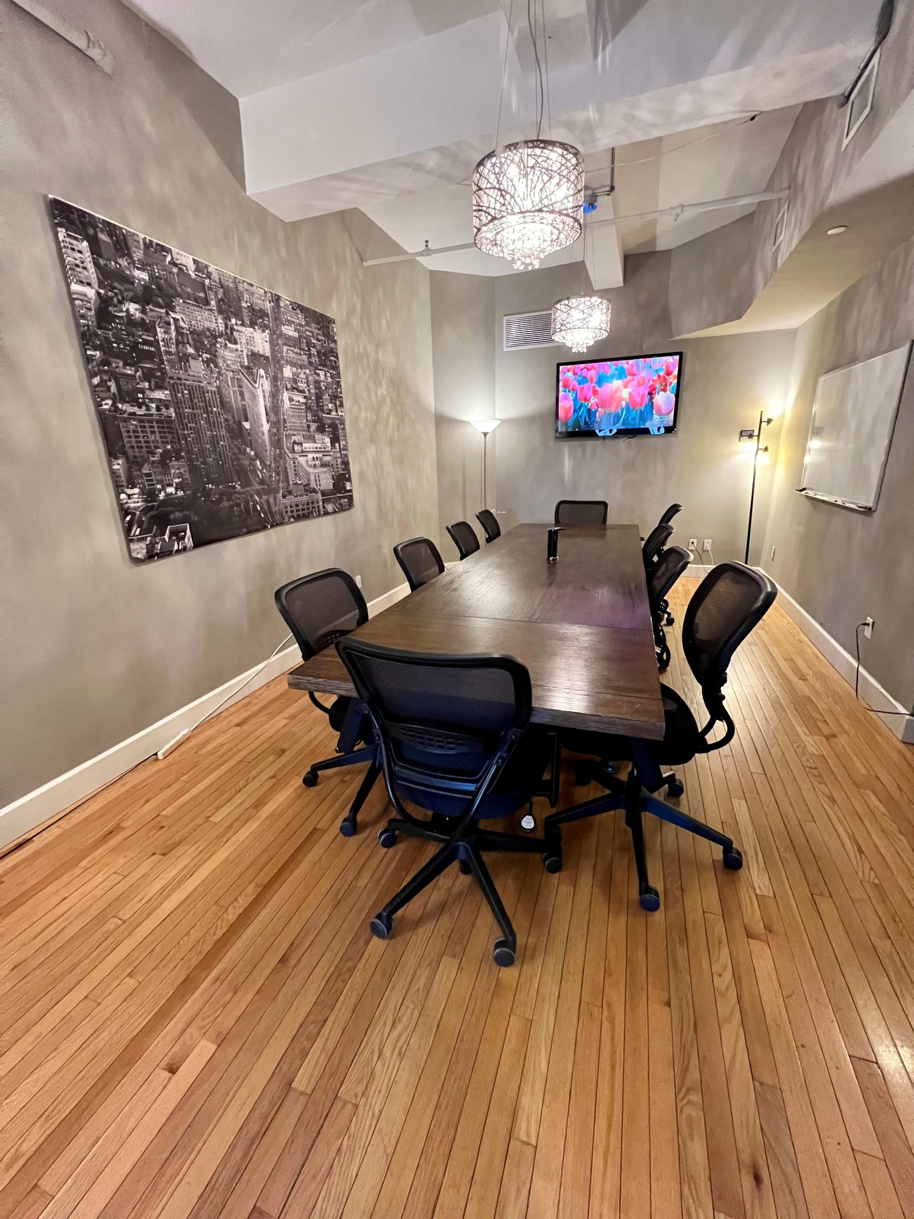 A long wooden conference table surrounded by ergonomic chairs is set in a well-lit meeting room featuring a large wall photo and a screen displaying vibrant images.