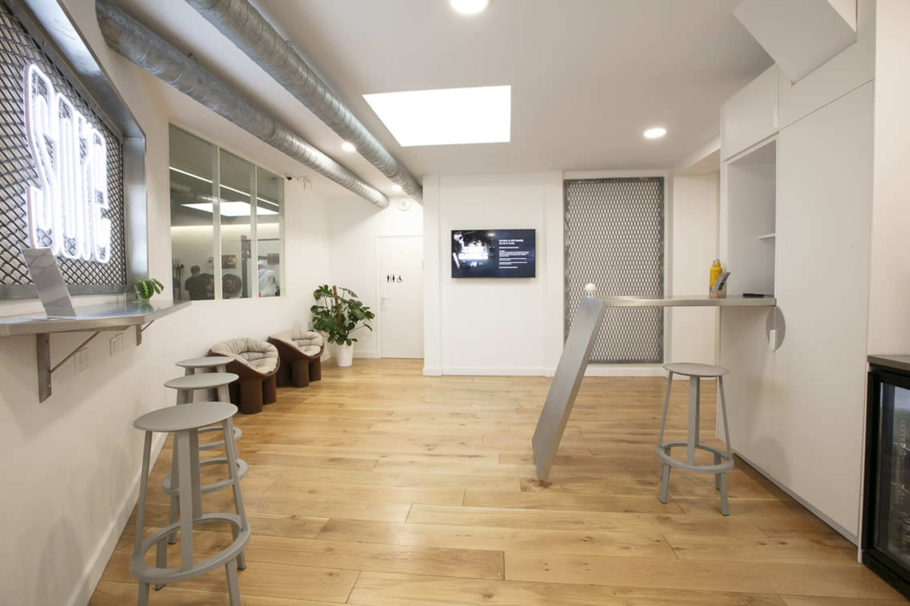 The interior of a modern room features a light wooden floor, a bar-style table with stools, and a television mounted on the wall.