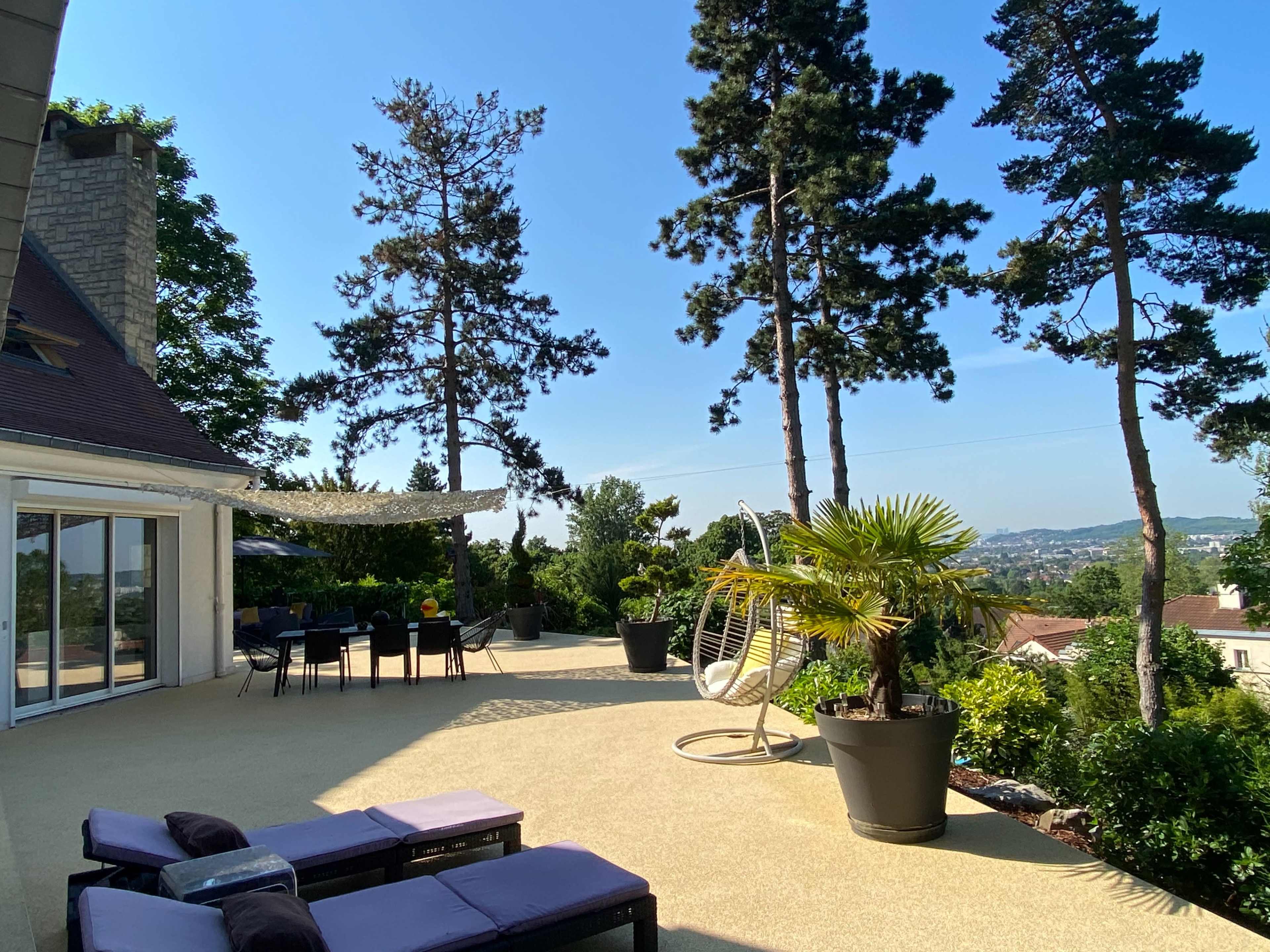 A spacious patio with lounge chairs, a hanging chair, and potted plants overlooks a scenic view surrounded by tall trees.
