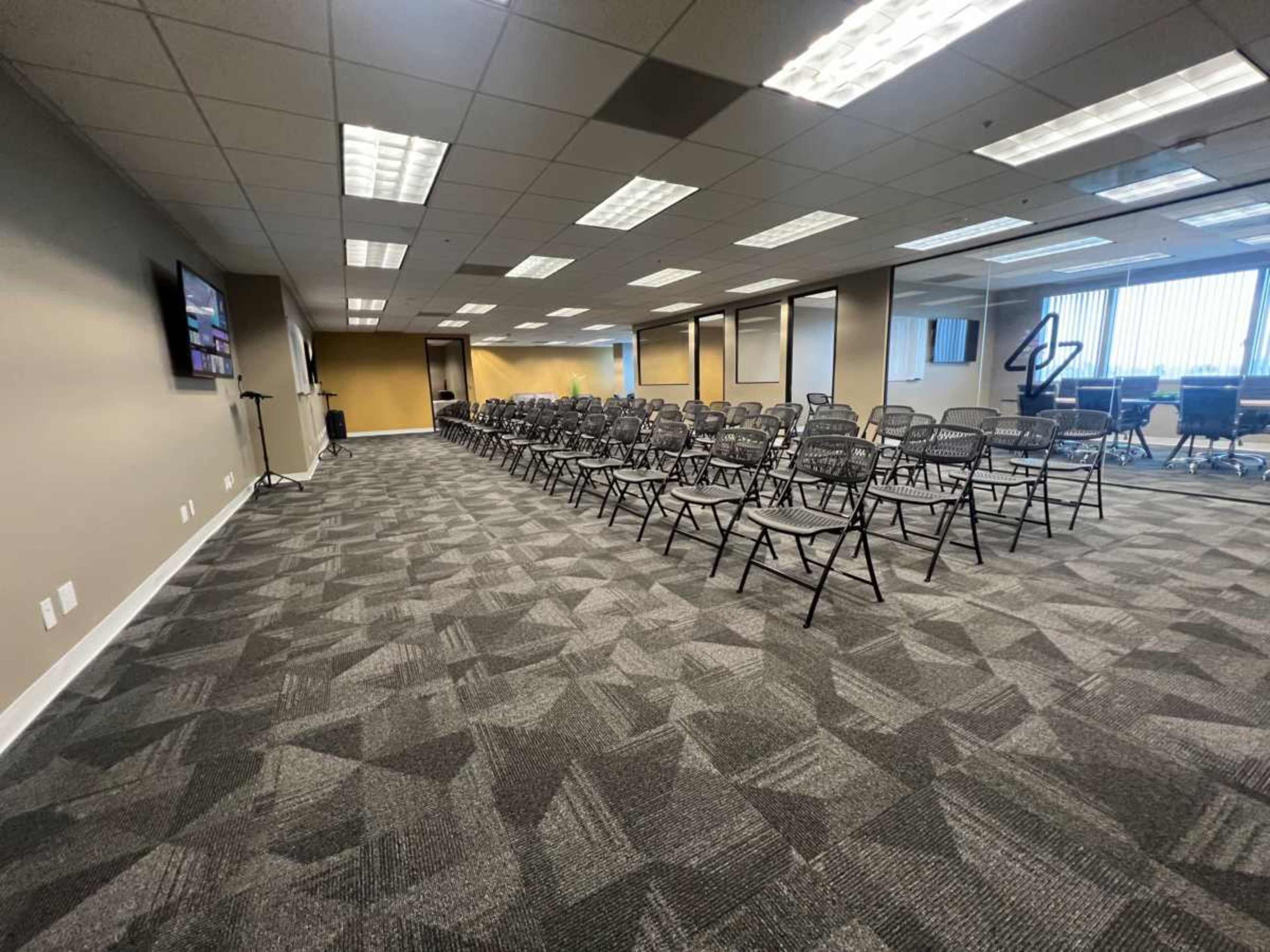 The image shows a conference room set up with rows of folding chairs facing a monitor on one wall.