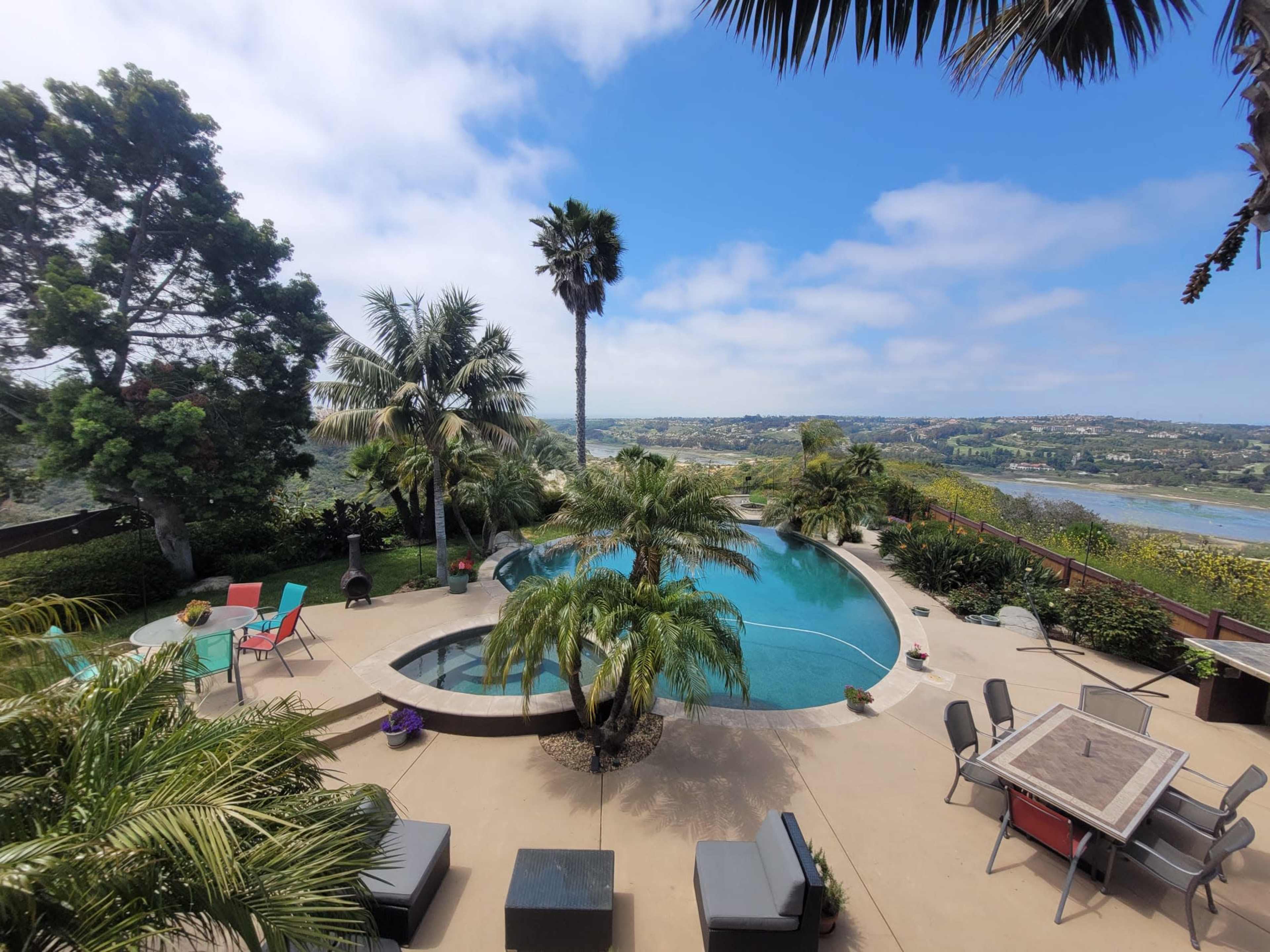 The image shows a residential backyard with a large pool surrounded by palm trees and patio furniture, overlooking a scenic view of rolling hills and a body of water.
