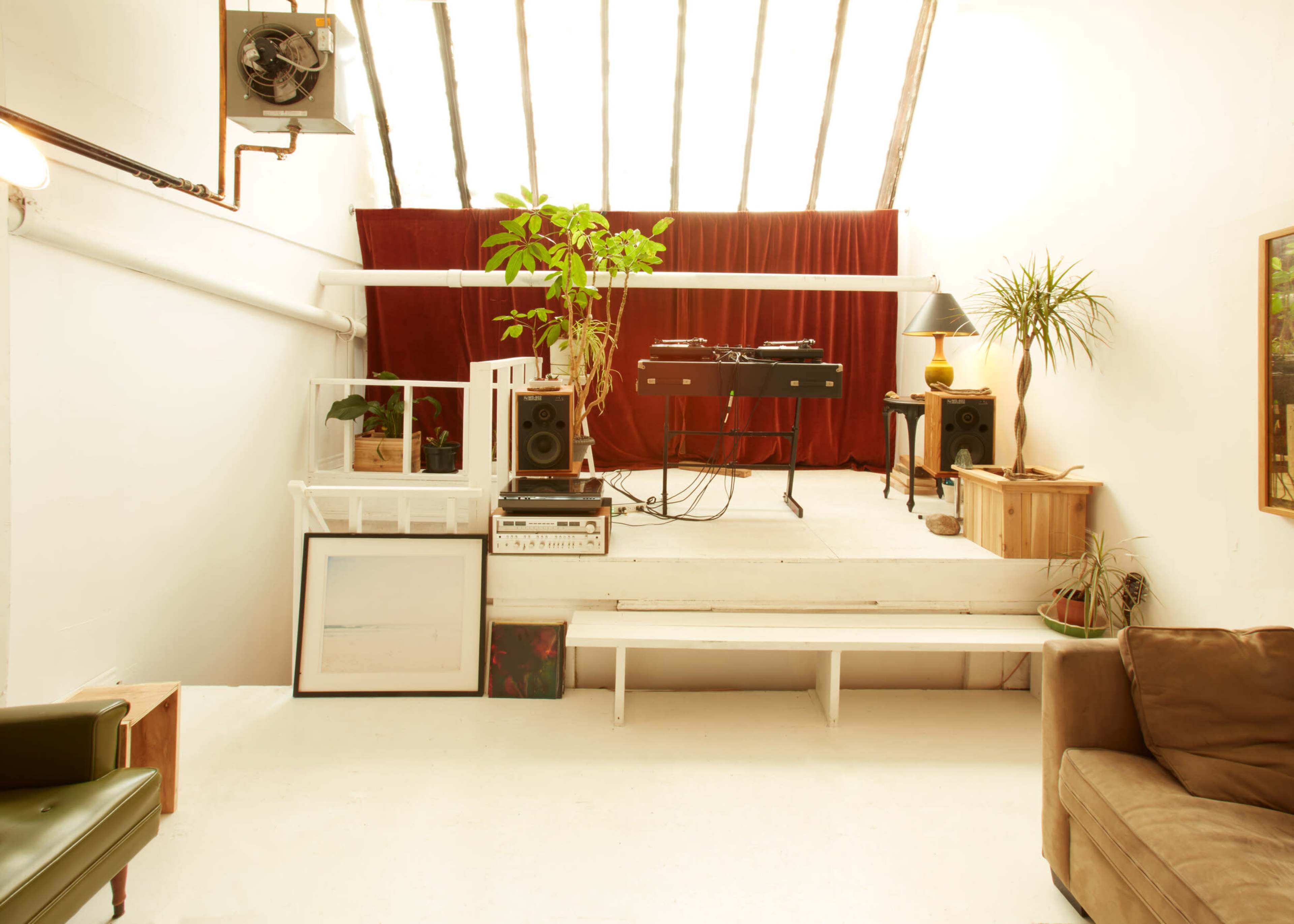 A bright, minimalistic interior with a raised platform featuring a DJ setup, plants, and various pieces of furniture against a backdrop of red curtains.