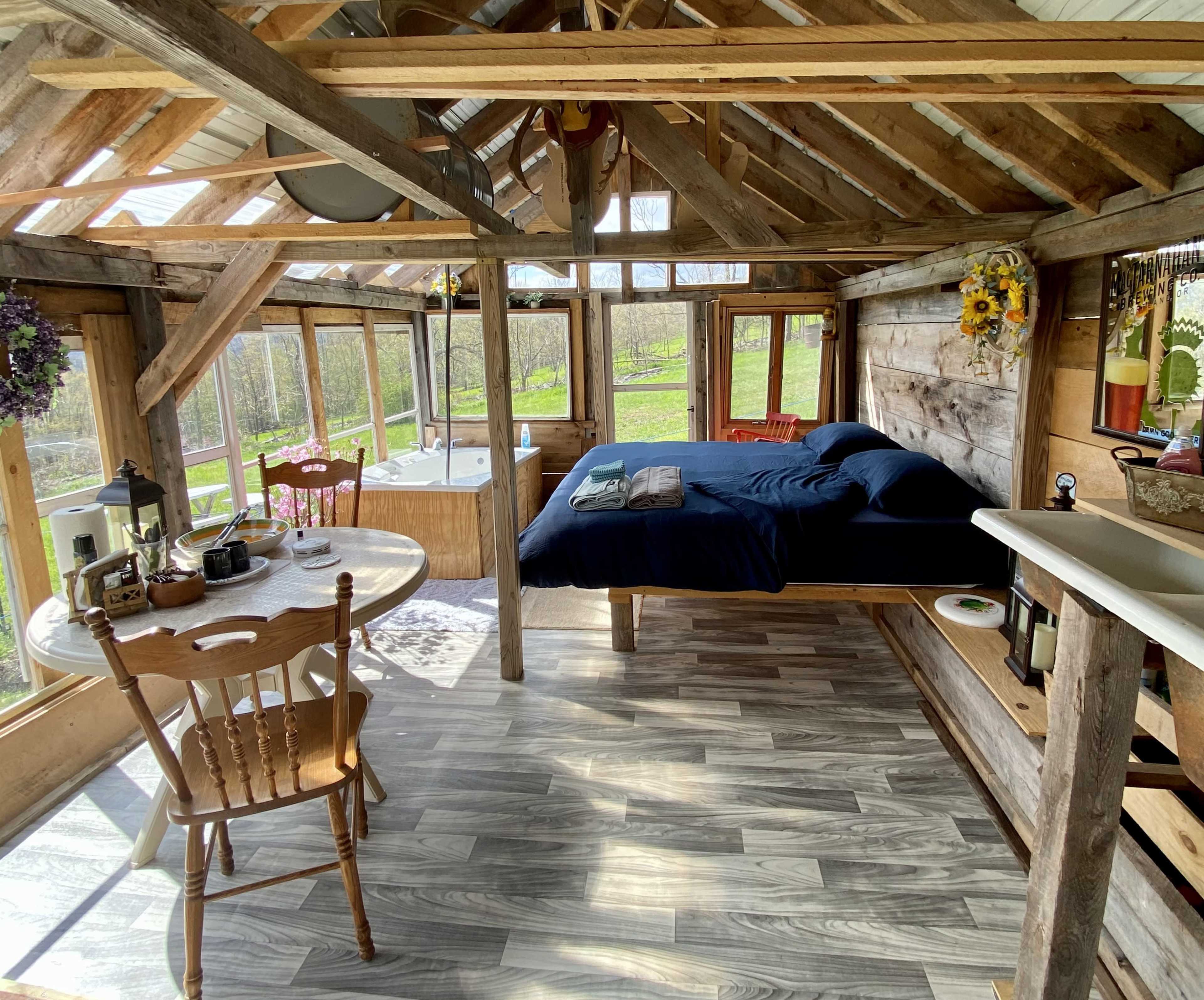 A rustic wooden cabin interior featuring a bed, a dining table with chairs, and a large window area overlooking a grassy landscape.