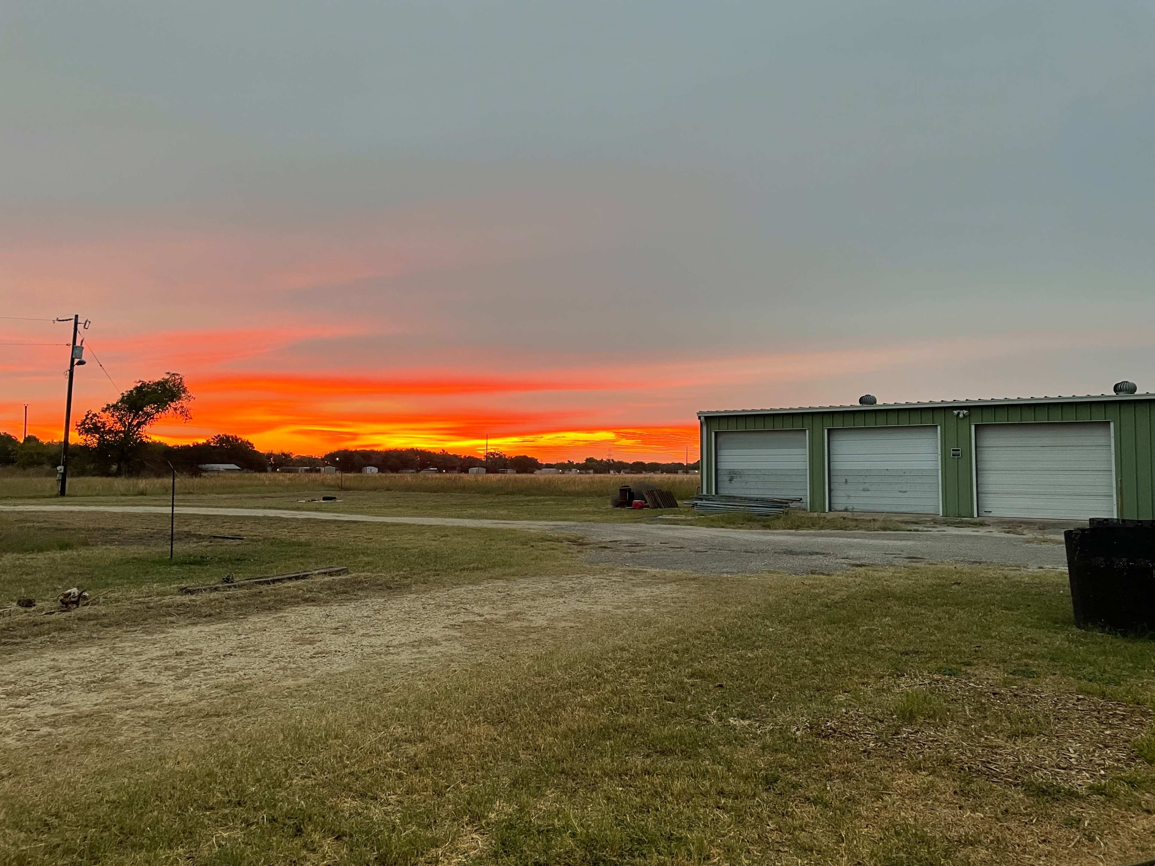 A vibrant sunset casts hues of orange and pink over a grassy field beside a green storage building with closed garage doors.
