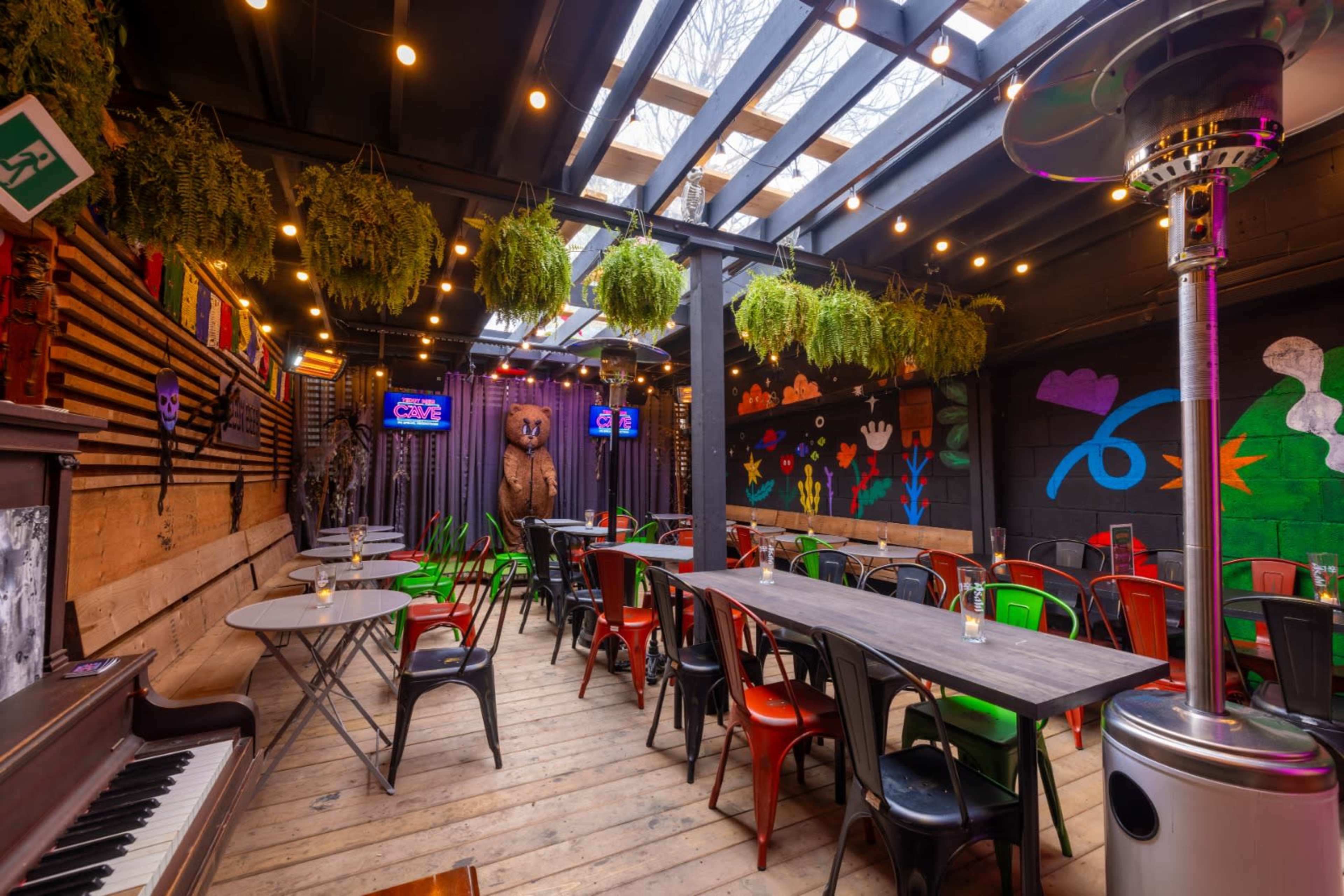 The image shows a vibrant restaurant interior with wooden floors, colorful chairs arranged around tables, and greenery hanging from the ceiling under a transparent roof.