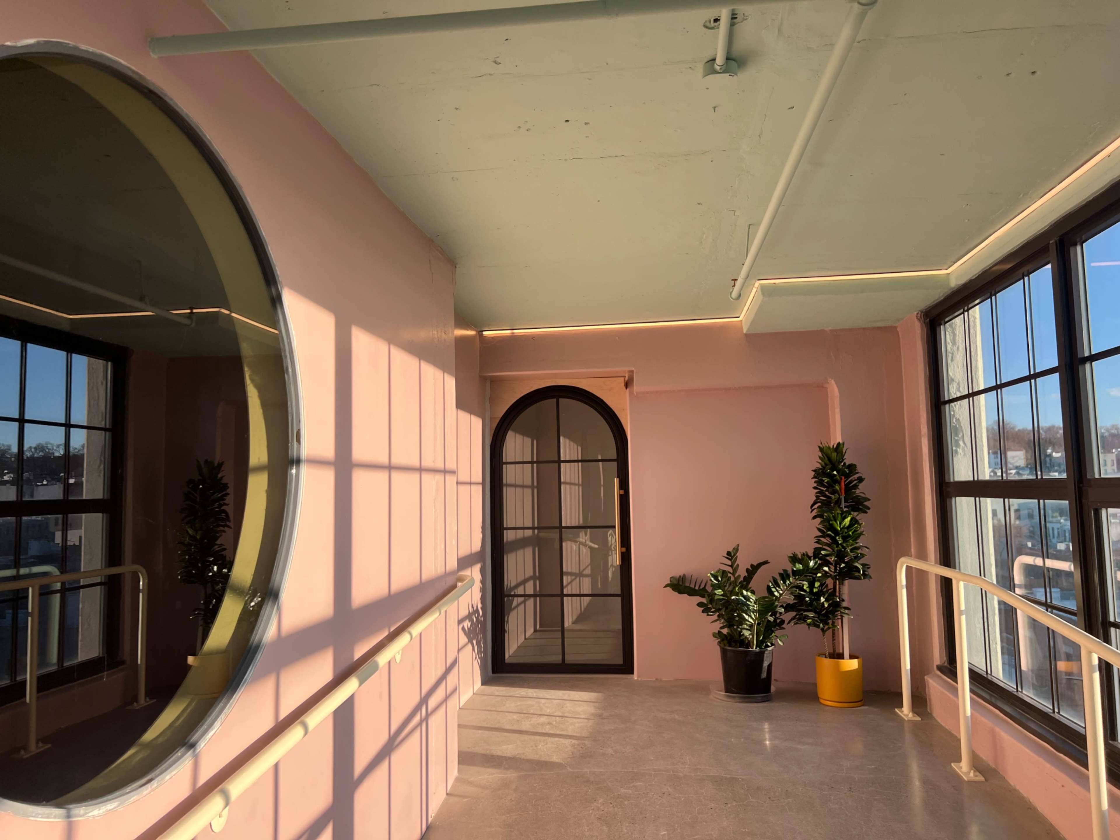 The image shows a brightly lit corridor with pink walls, large windows, and an arched doorway leading to another space, along with potted plants in two corners.