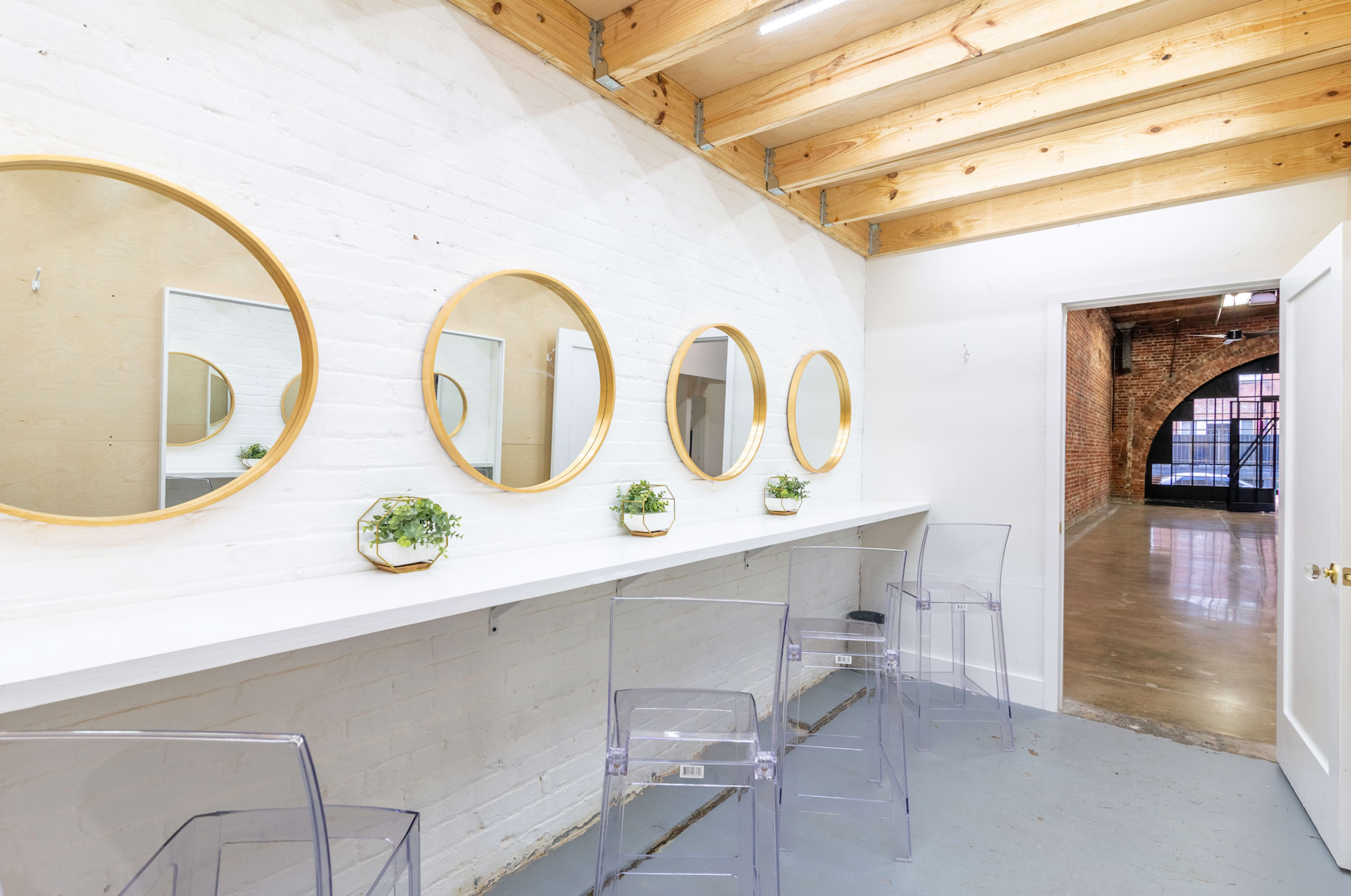 A modern, minimalist room with a row of five circular mirrors above a white countertop and clear acrylic chairs, leading into a space with exposed brick walls.