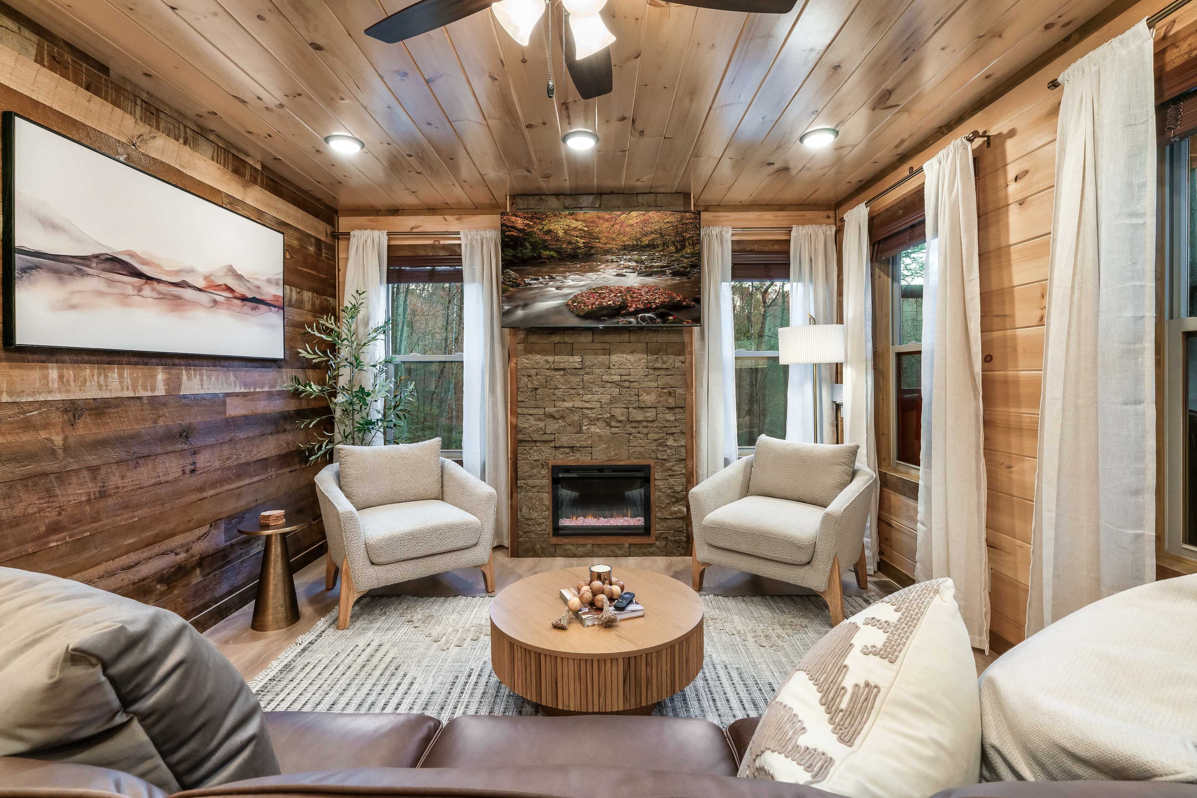 The image shows a cozy living room with wooden walls, two armchairs facing a circular coffee table, and a fireplace framed by large windows with sheer curtains.