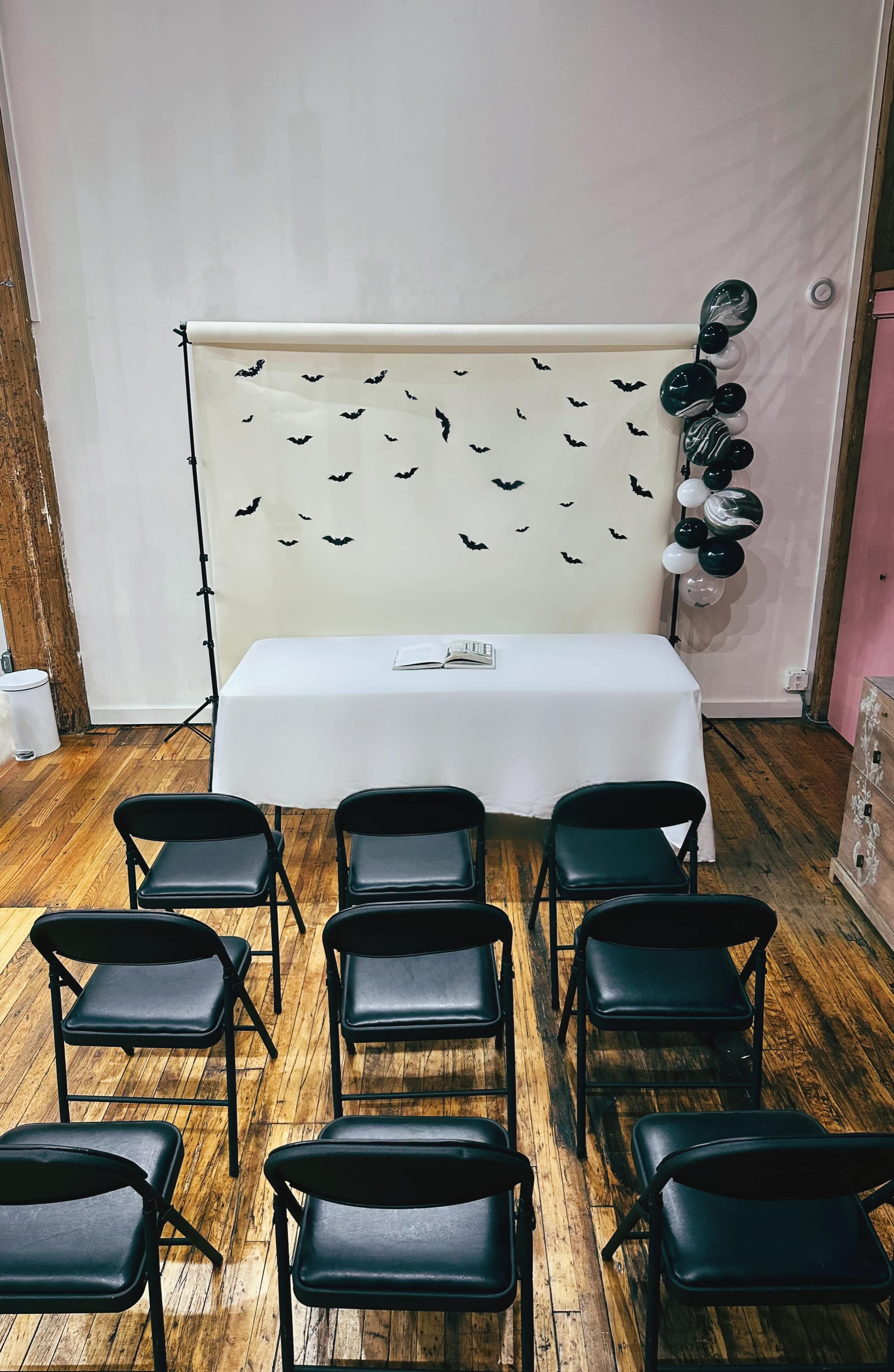 The image shows a room with a neatly arranged row of black folding chairs facing a table draped in white, set against a backdrop decorated with black bats.